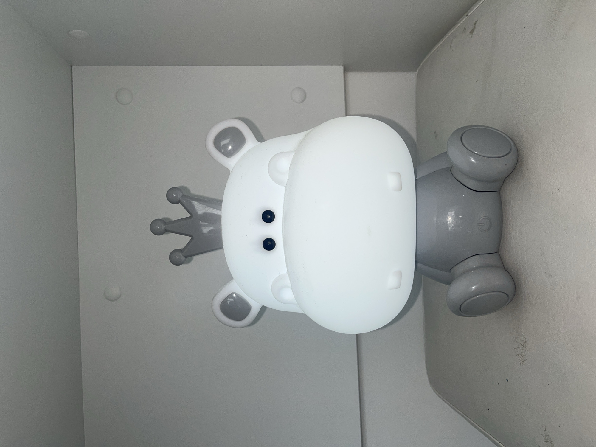 The image shows a gray and white hippo-shaped lamp with a crown on its head. The lamp is inside a white cupboard or shelf. The location is unknown and cannot be determined from the image.