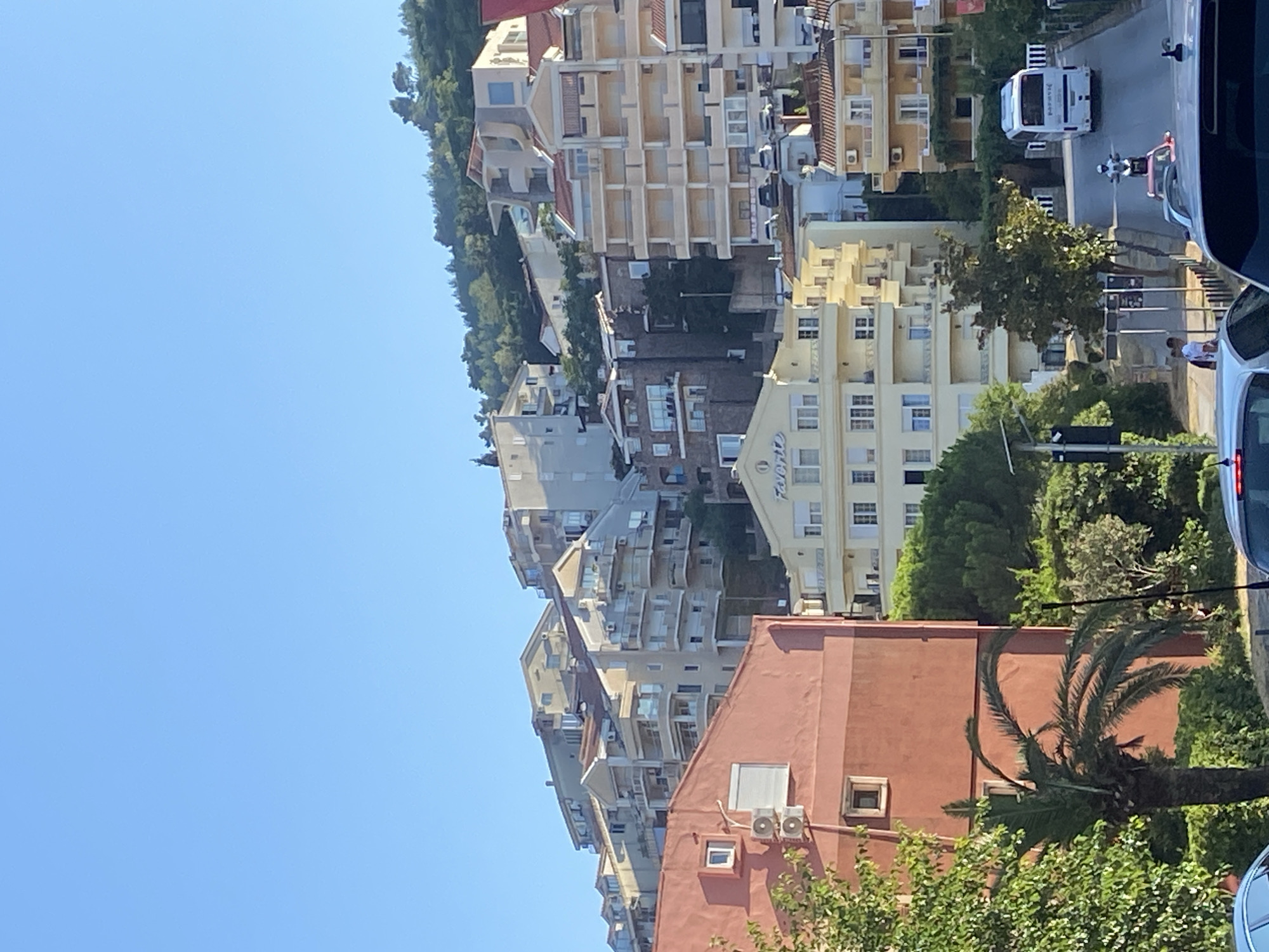 The image shows a sunny day in the city of Budva, Montenegro. Buildings are constructed on a hillside, with a clear blue sky above. In the foreground, there are several cars on a street, and a motorcyclist is riding a white motorcycle. A white bus is visible further up the street. There are trees and palm trees in the scene. One building has the word "Favorit" written on it in a script font. The buildings are multi-story structures with balconies.