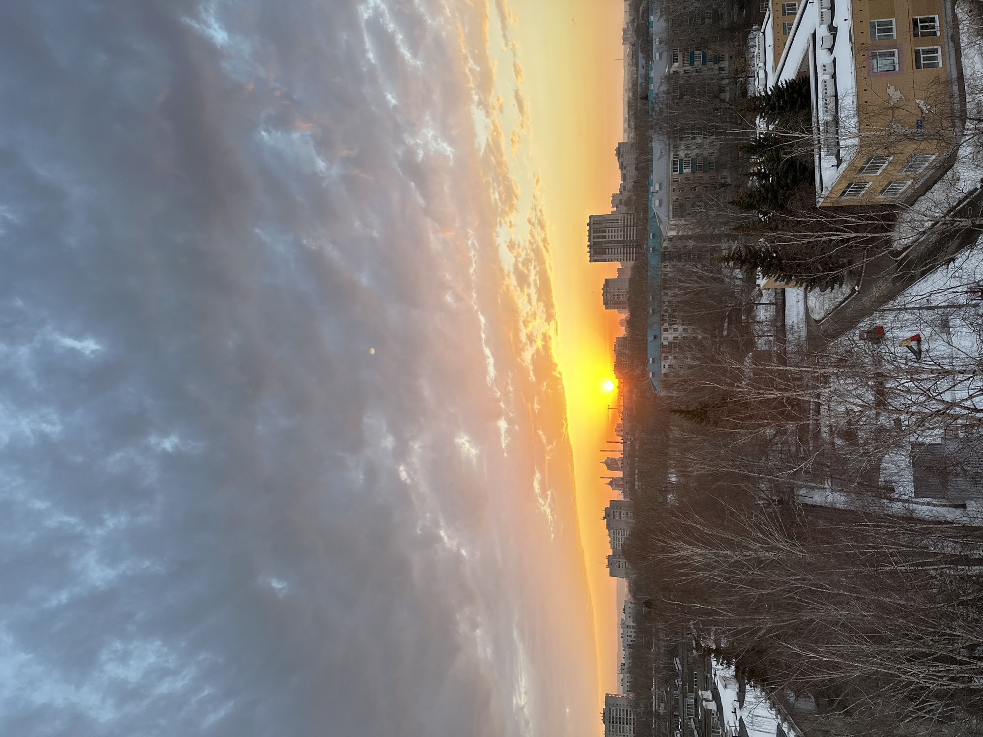 The image depicts a winter sunset in Kazan, Russia, viewed from an elevated position overlooking a cityscape. The sun is setting in the distance, casting a bright orange and yellow glow across the sky. To the left, heavy gray clouds fill a significant portion of the sky. The cityscape is composed of residential buildings of varying heights, and bare deciduous trees are visible in the foreground. Snow covers the ground and rooftops. The general atmosphere of the scene is cold and urban, with the warm sunset contrasting with the winter environment.