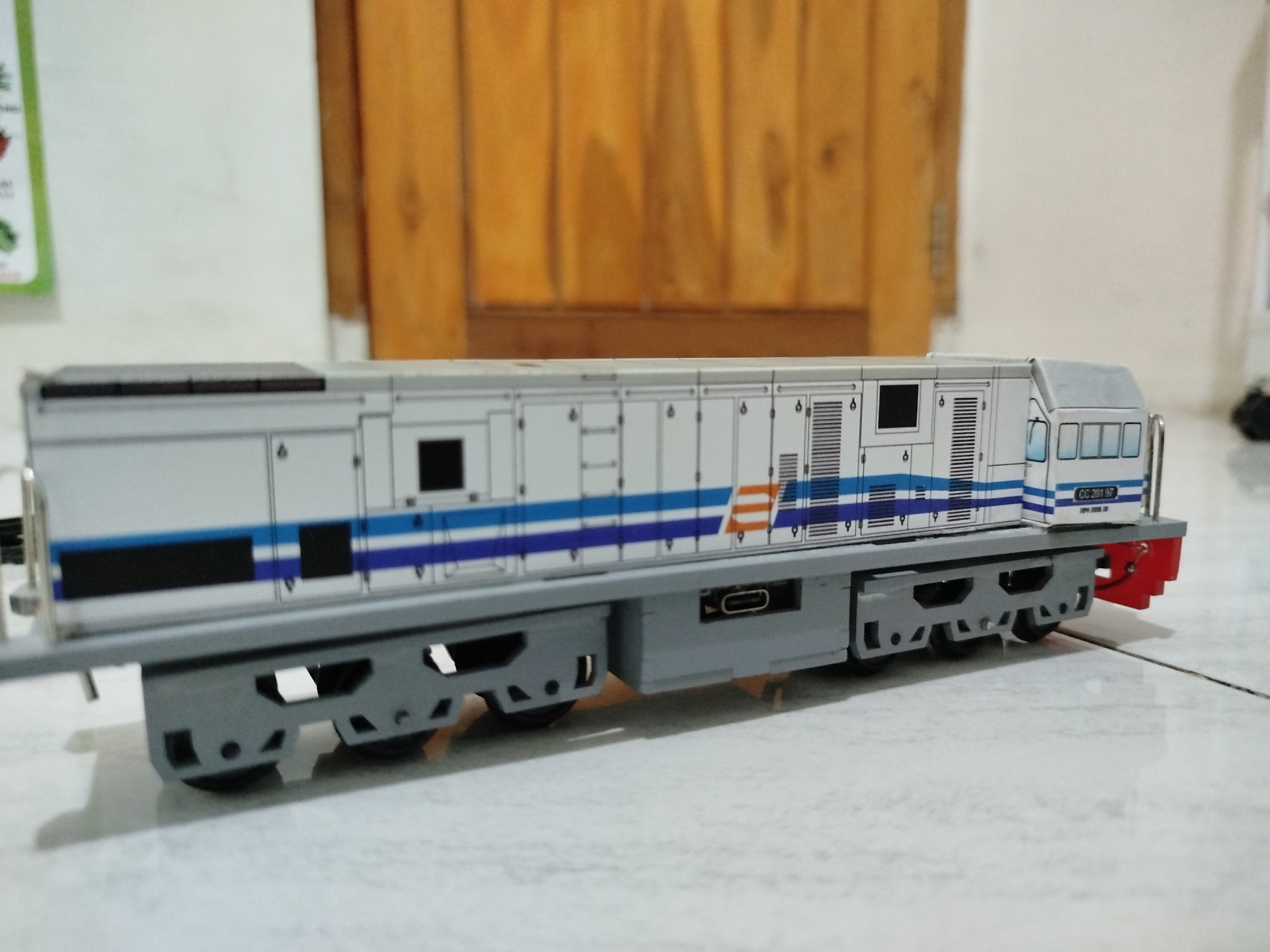 A close-up, eye-level, medium shot captures a model train engine on a white tiled floor. The engine is primarily white with blue and orange stripes. It has detailed features like windows, doors, vents, and a small red piece at the rear. The train is angled towards the right side of the frame. The background is softly blurred, showing a wooden surface and a white wall, suggesting an indoor setting. The lighting is moderate, with shadows cast on the floor. No people are present in the image. The location is not specified in the image or the prompt.