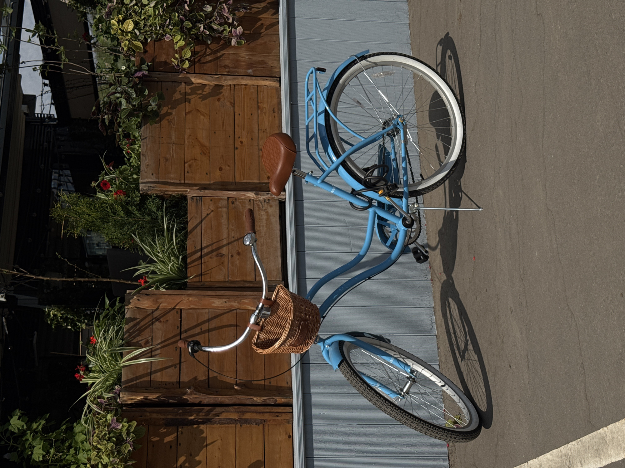 A light blue bicycle with a wicker basket on the handlebars is parked next to a wooden wall or fence. The seat is brown leather. The bicycle is on a gray pavement. A planter with greenery and red flowers is on top of the wall/fence. The location is unknown; there is no indication this is in Moscow, Russia.