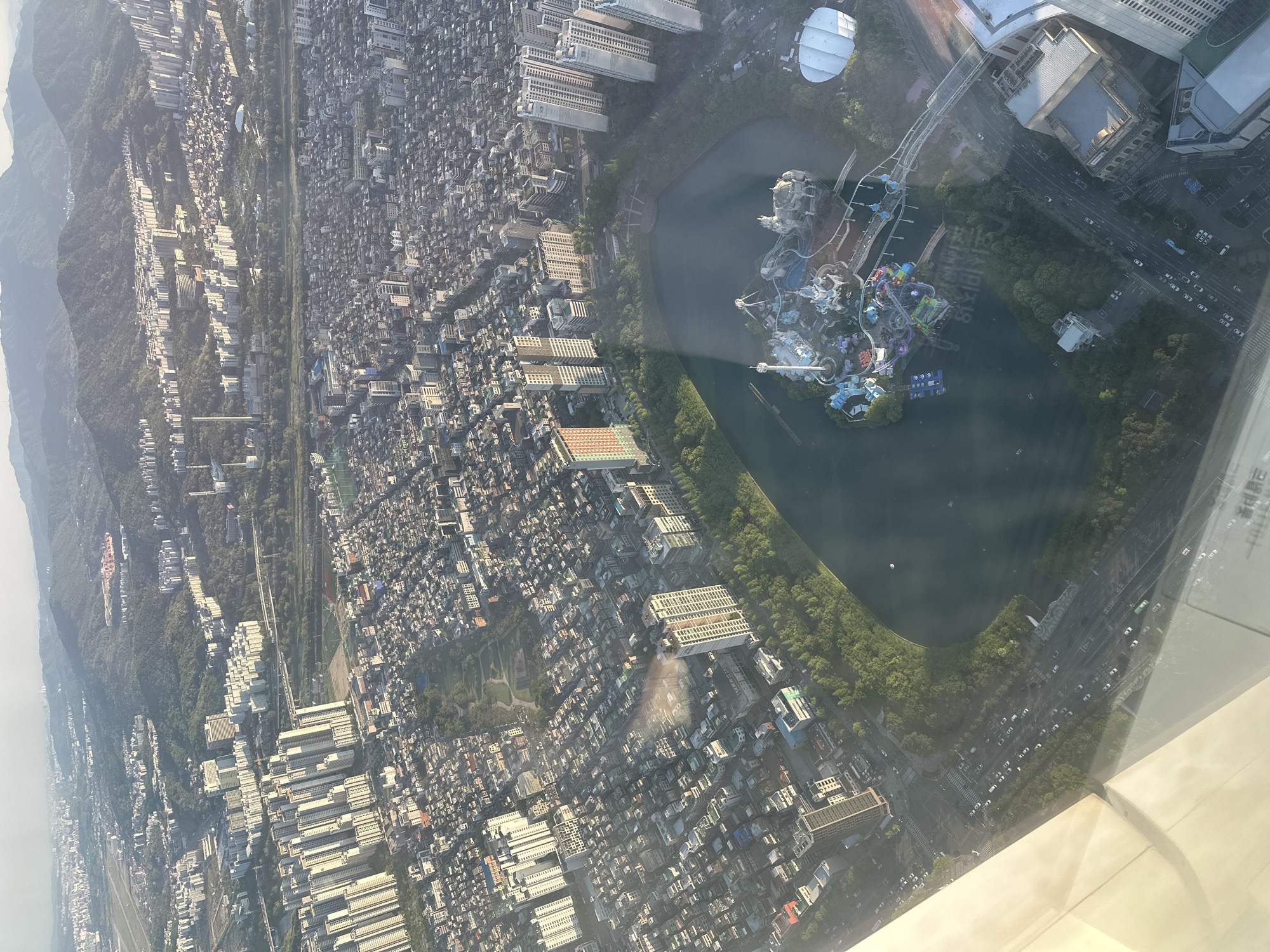 The image shows an aerial view of Seoul, South Korea, specifically the area around Seokchon Lake. The lake is visible in the center, surrounded by lush greenery and adjacent to the Lotte World amusement park, which is partially visible within the lake. The park features distinctive structures and rides. Dense urban development surrounds the lake and park, including residential buildings, commercial areas, and roadways. Mountains are faintly visible in the background. The photograph appears to be taken from a high vantage point, likely from a plane or tall building. The bottom right corner of the image shows a part of the window from which the picture was taken.