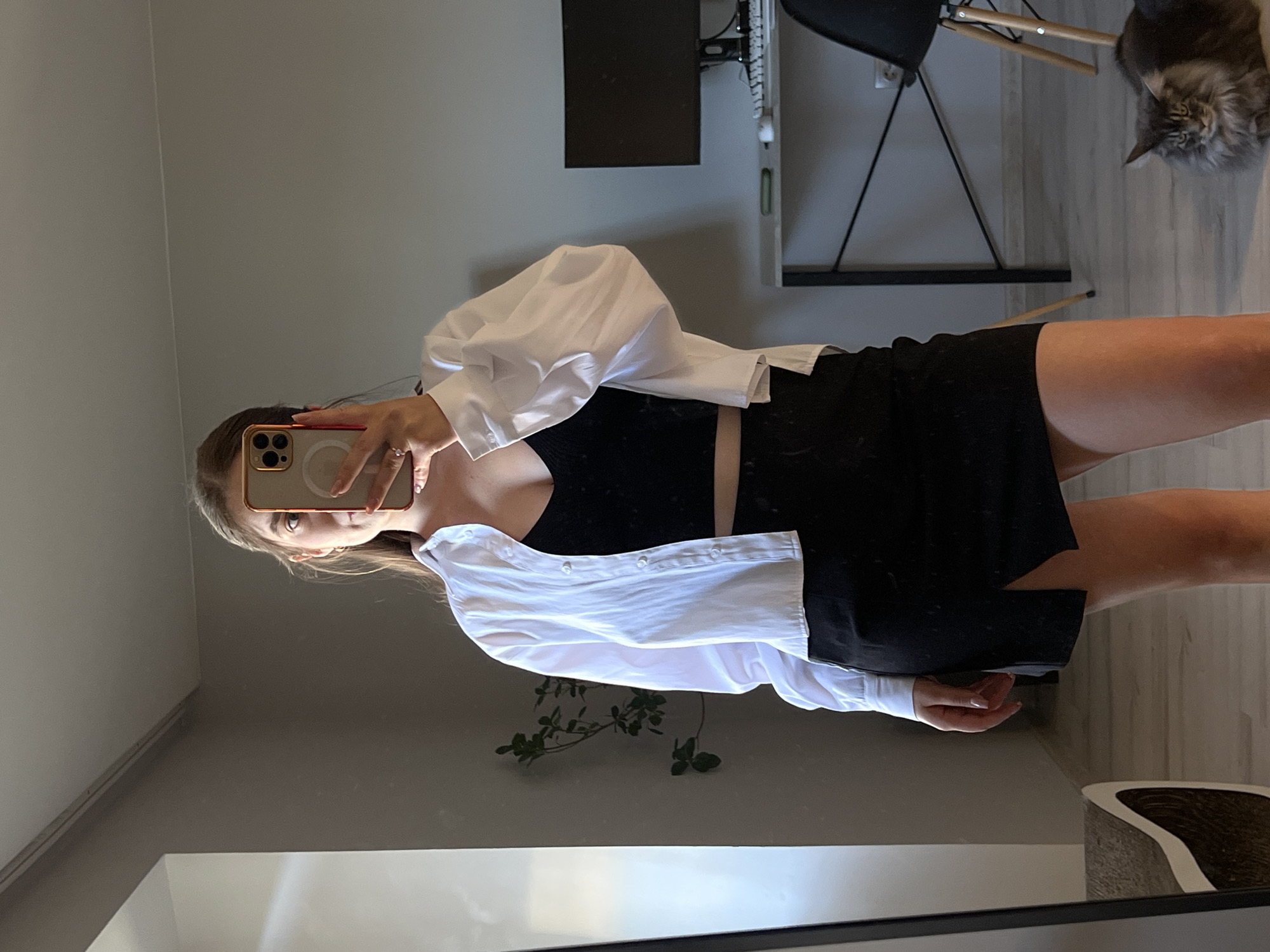 A young woman with long brown hair stands in a room, taking a mirror selfie with her phone. She is wearing a white, unbuttoned shirt over a black crop top and a black skirt with a slit. The room appears to be a home office, with a desk, computer monitor, and chair visible in the background. A cat is lying on the floor to the right of the frame. There is no visible indication that this image was taken in Ternopil, Ukraine, or anywhere else.