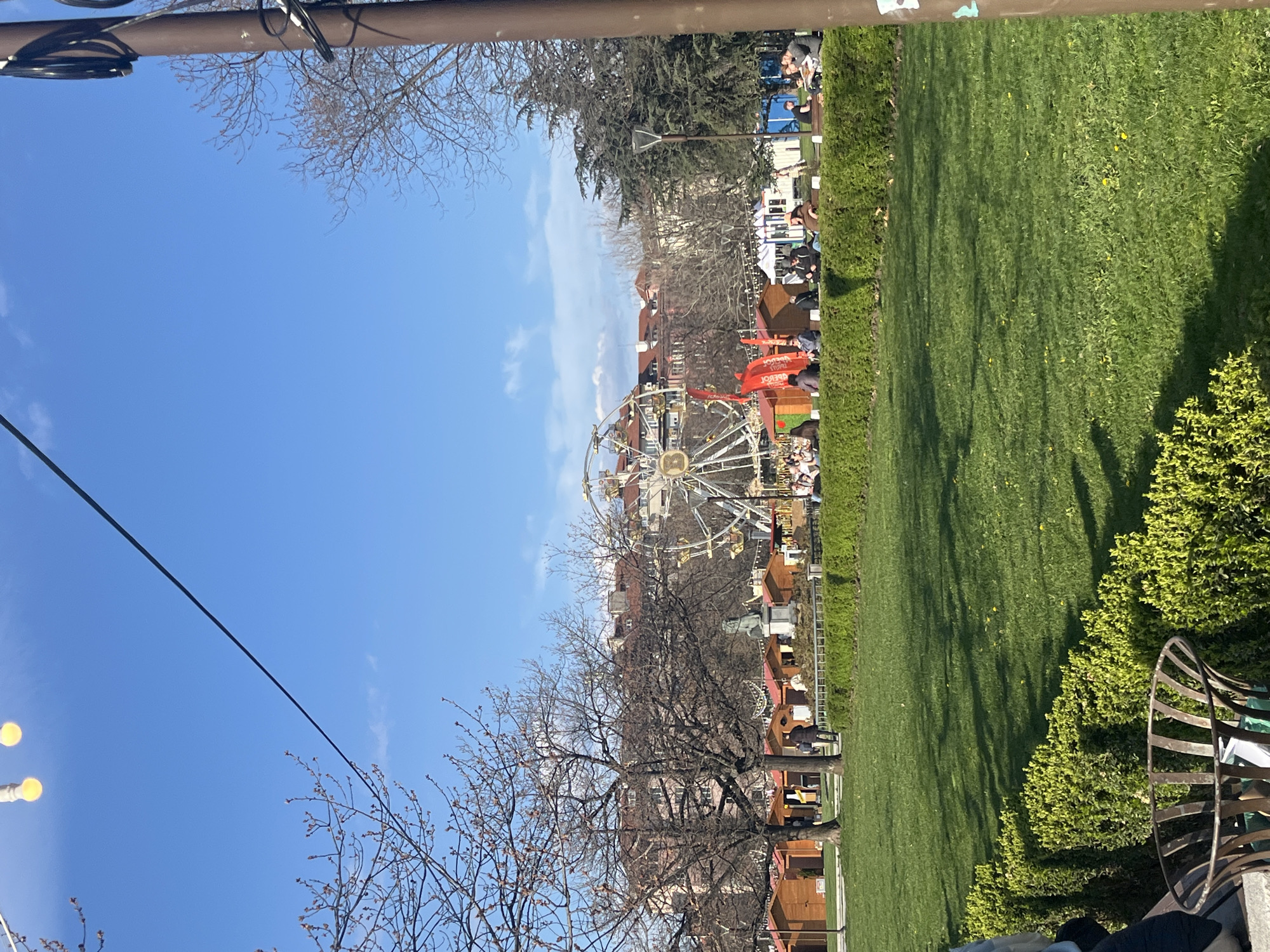 The image shows a park setting in Sofia, Bulgaria, likely the Christmas market at the City Garden. A Ferris wheel stands prominently amidst buildings, trees, and market stalls. People are seen in the background. The foreground features a grassy area, bushes, a partially visible bench, and bare trees. The sky is a clear blue with some clouds. A utility pole with wires is visible at the top and left of the frame.
