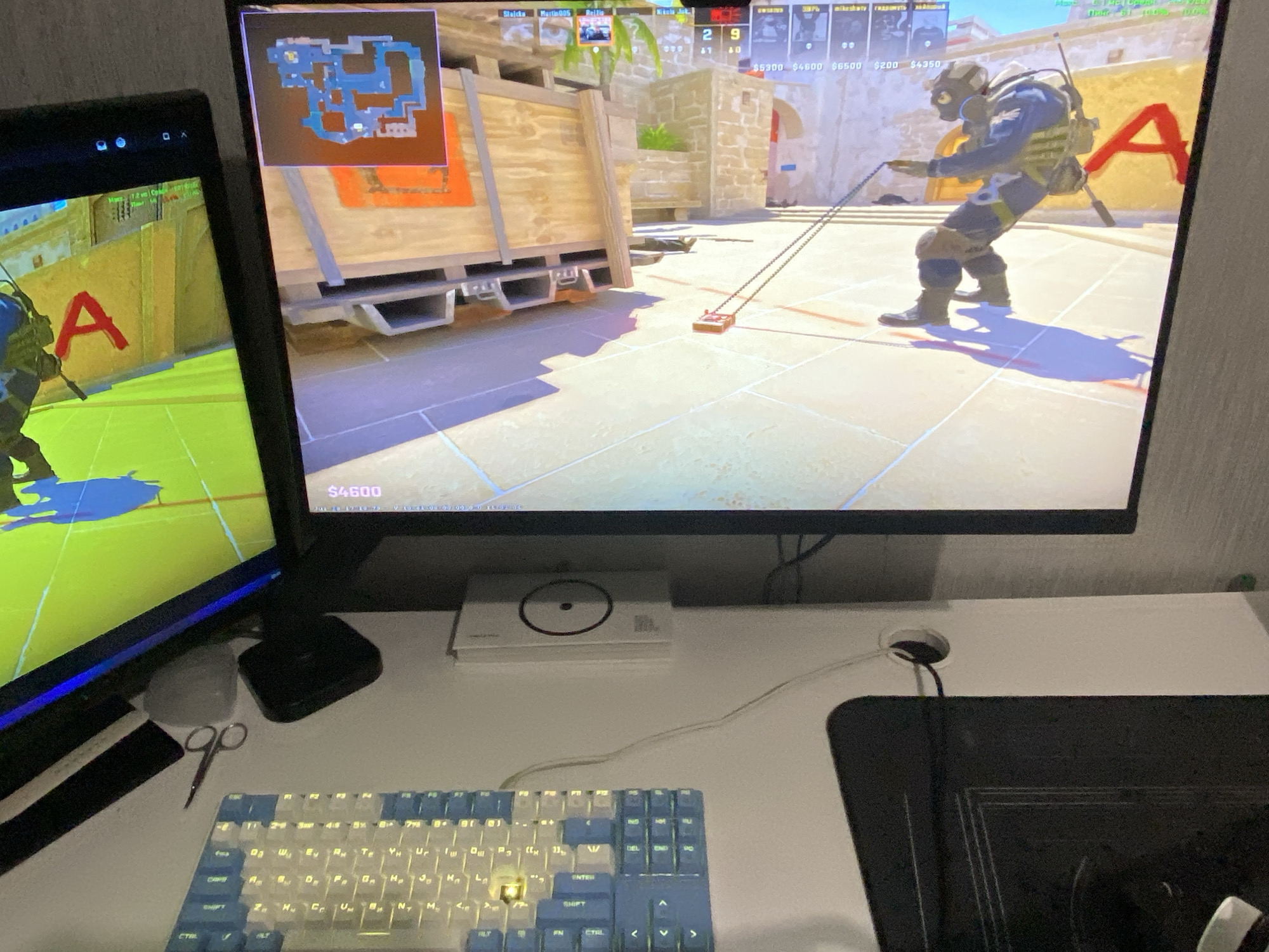 The image shows a gaming setup with two computer monitors displaying the video game Counter-Strike: Global Offensive. The main monitor on the right shows a player character holding a defusal kit. The monitor on the left displays a portion of the game's map. In the foreground, a white and blue mechanical keyboard is visible. A pair of scissors and a small, grey object, possibly a mouse, are on the desk to the left of the keyboard. To the right of the keyboard, a black mousepad and a hole in the desk for cable management are present. A white rectangular object with a black circle in the center is situated on the desk below the main monitor. The setting appears to be indoors, likely a room with a textured grey wall in the background. There is no information in the image that would indicate the location as Saratov, Russia.