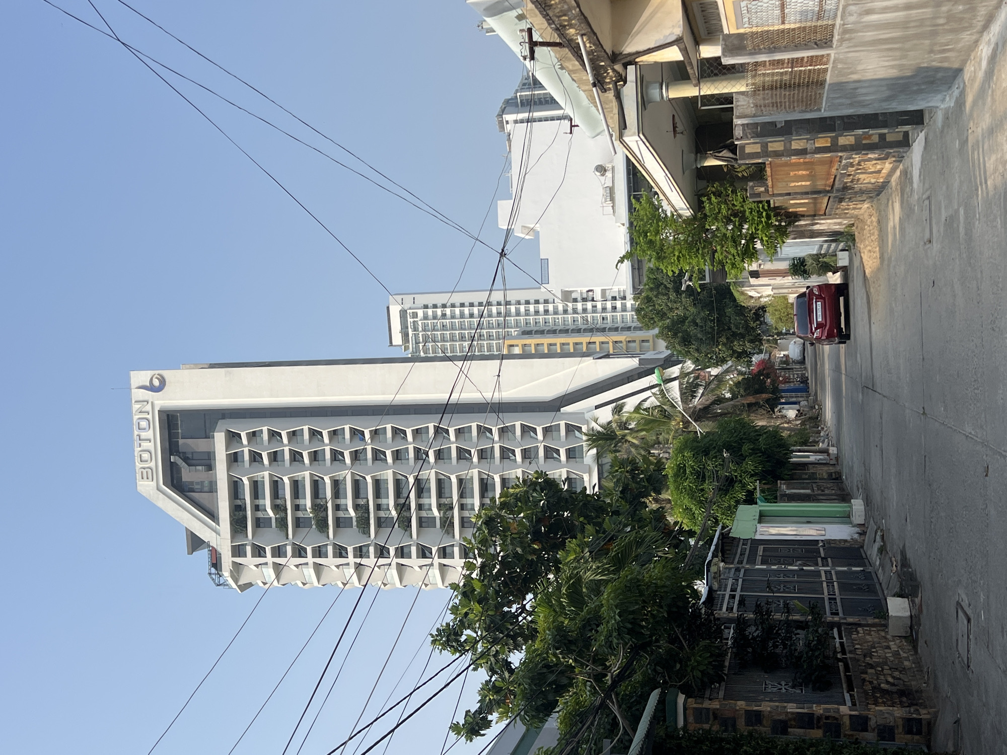 A ground-level shot, rotated counter-clockwise, depicts a street scene in what is likely Bắc Nha Trang, Vietnam. A tall, modern, white hotel building is visible on the left with the name "BOTON" displayed vertically. The hotel has a honeycomb-like facade. Overhead power lines crisscross the view. On the right side of the image is a street lined with various buildings, mostly residential, and trees. A red car is parked down the street. The sky is clear and blue.