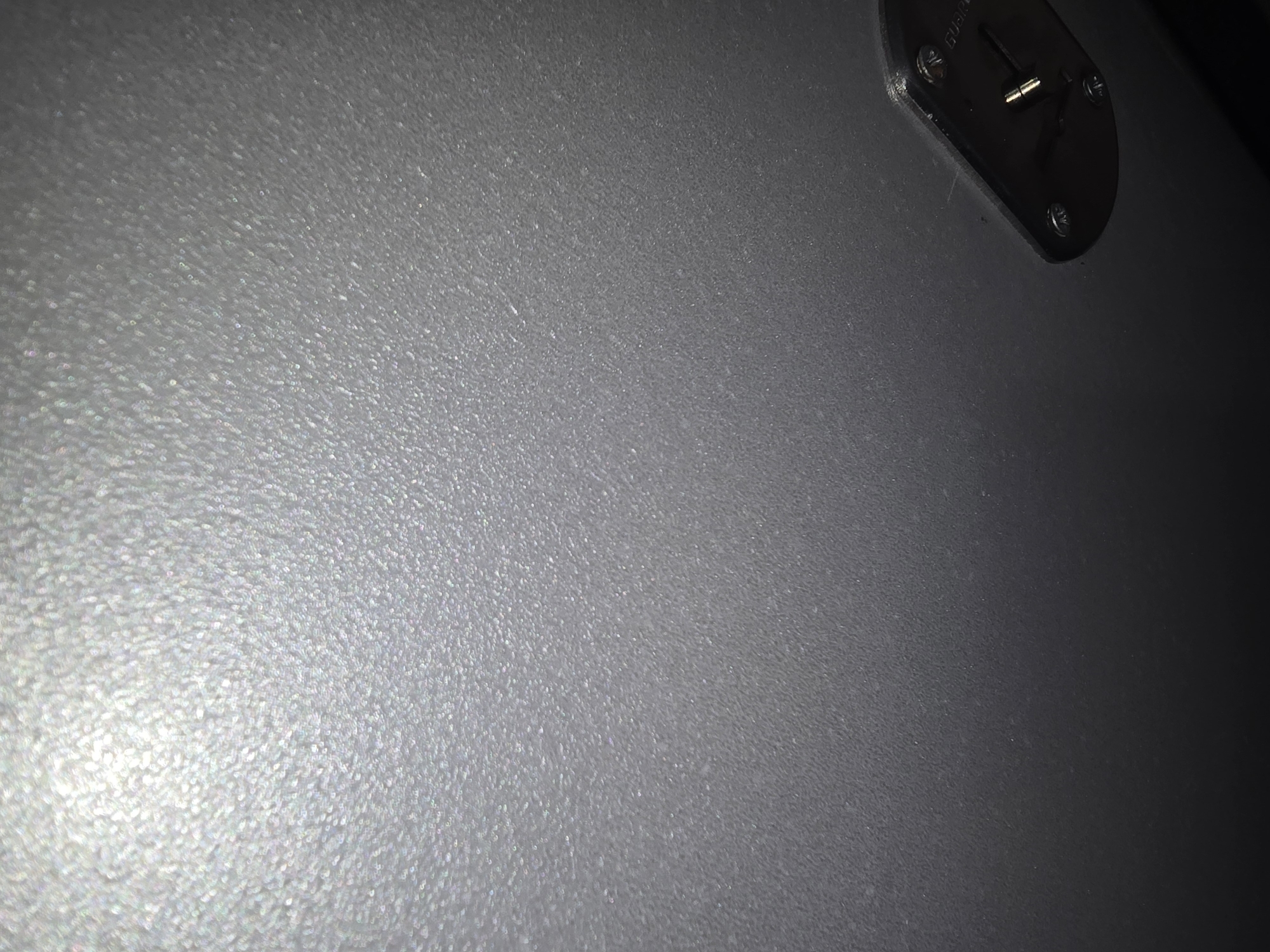 The image shows a textured, metallic surface, likely the interior of a vehicle or a piece of equipment. A metallic fixture, possibly a lock or a latch, is visible in the lower right corner. The surface is a neutral gray with a speckled texture, reflecting light in a way that creates a shimmering effect. The lighting suggests the image was taken indoors or in a dimly lit environment, with a bright light source creating a strong highlight on the upper left portion of the surface. There are no people or visible signs of the city of Ulan-Ude, Russia.