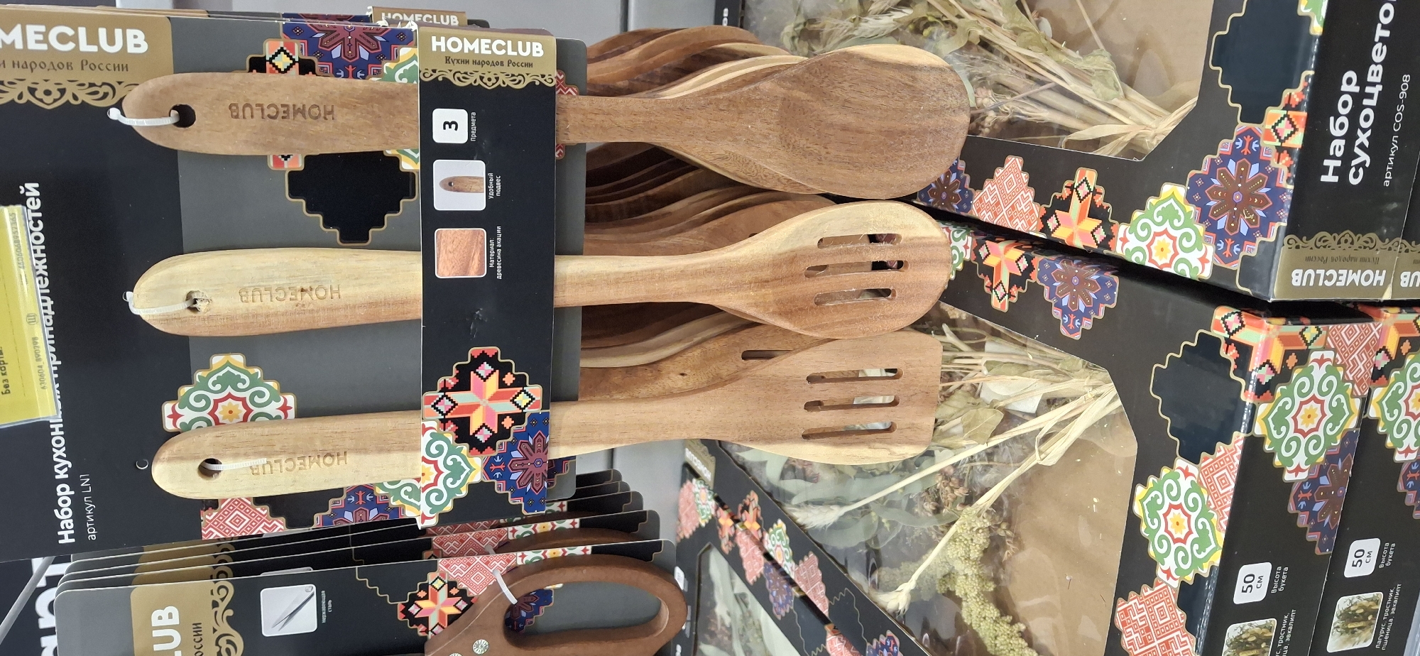 The image shows a close-up view of a retail display of wooden kitchen utensils, specifically spatulas and a slotted spoon, with ornate packaging. The utensils are made of light-colored wood, and some have "HOMECLUB" branded into the handles. The packaging features a black background adorned with colorful, geometric folk-art patterns in red, green, blue, and yellow. The text on the packaging is in Russian and indicates that it is a "Set of kitchen utensils" with the article number LN1 and "HOMECLUB" branding. There is also a mention of "wood of acacia" and "convenient hanging loop." Further text on the packaging includes "3 предмета" (3 items). In the background, more of the same type of kitchen utensil sets are visible, as well as what appears to be dried floral arrangements or bouquets packaged in boxes with similar folk-art designs. The items are arranged vertically in what seems to be a retail store setting. The image is taken from a slightly low angle, looking up at the display. The location of Тельмана, Russia, is not visible in the image and cannot be determined from the visual information provided.