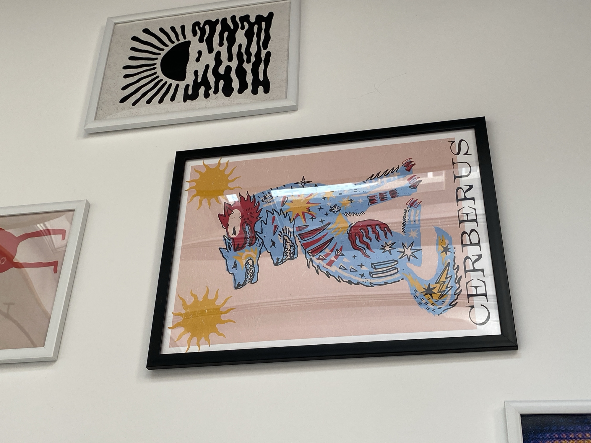 The image shows a section of a white wall adorned with several framed art pieces. At the top is a white-framed print with a black abstract design resembling a sun and dripping shapes. Below it and to the right is a black-framed picture featuring an artistic representation of Cerberus. The artwork has a light pink background and depicts a three-headed dog with a blue patterned body, with the word "CERBERUS" written vertically along the right side. To the left is another white-framed piece with a minimalist human figure in a bright color. In the bottom right corner, a small part of another framed picture is visible. The scene is generally well-lit. There is no indication of this scene being in Izhevsk, Russia.