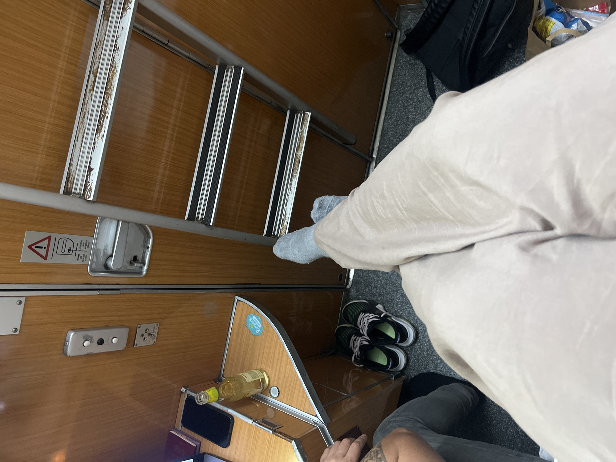 The image depicts the interior of a train compartment, likely a couchette or sleeping car, but location context is irrelevant. A ladder extends from the top bunk. A partially visible figure is seated in the compartment, wearing light-colored trousers and grey socks. A pair of running shoes is on the floor. A bottle of liquor rests on a small shelf. The overall condition of the train's interior is worn, with visible scratches and aging on the wood panels and metal fixtures. The picture includes a black bag and a few personal items.