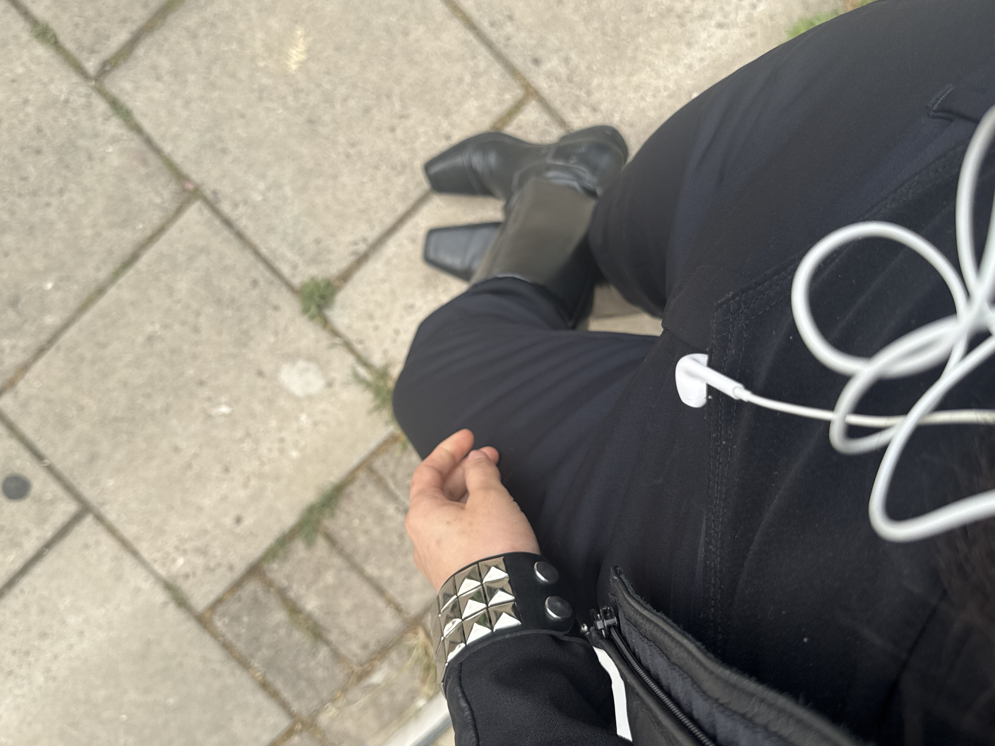 The image shows a person sitting outdoors, presumably on a ledge or low wall, based on the visible pavement tiles. The person is wearing black trousers, black boots, and a black jacket. A studded wristband is visible on their left wrist. White earphones are tangled and attached to a device placed in the pocket. The pavement is made of light grey square tiles. It is not possible to ascertain the exact location in London or any actions, interactions, or additional subjects without more context.