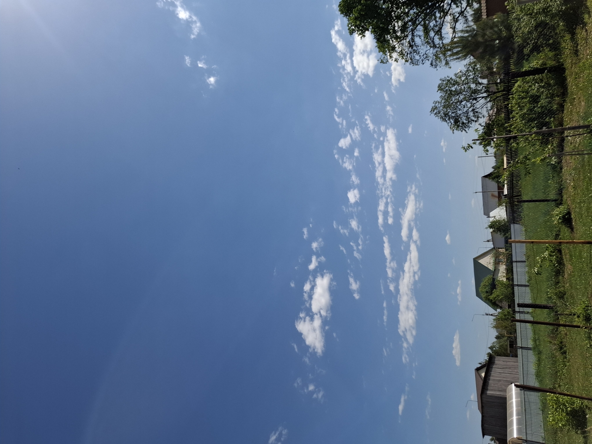 The image depicts a rural scene in Амзя, Russia, under a bright blue sky dotted with scattered white clouds. The horizon line features residential structures, including houses with pointed roofs and wooden outbuildings, partially obscured by a chain-link fence and overgrown greenery. Tall trees and bushes are interspersed along the property lines. Wooden poles are placed intermittently along the fence line. The foreground is dominated by the sky, with a gradient transitioning from a darker blue towards the top to a lighter shade closer to the horizon.