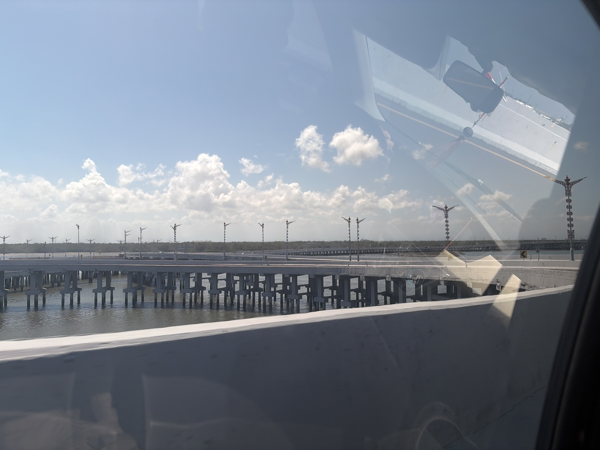 The image depicts a view of a bridge spanning over water, with a clear blue sky dotted with white clouds overhead. The bridge is characterized by its concrete structure supported by numerous pillars submerged in the water. The foreground shows a portion of a concrete barrier, possibly from a vehicle or another elevated structure. In the distance, the bridge continues, flanked by what appear to be streetlights or decorative poles. The reflection on the glass in the upper right corner shows the interior of a vehicle, including a rearview mirror with a dangling ornament. The scene is captured from the perspective of someone inside a vehicle looking out. The environment appears to be coastal or near a large body of water.