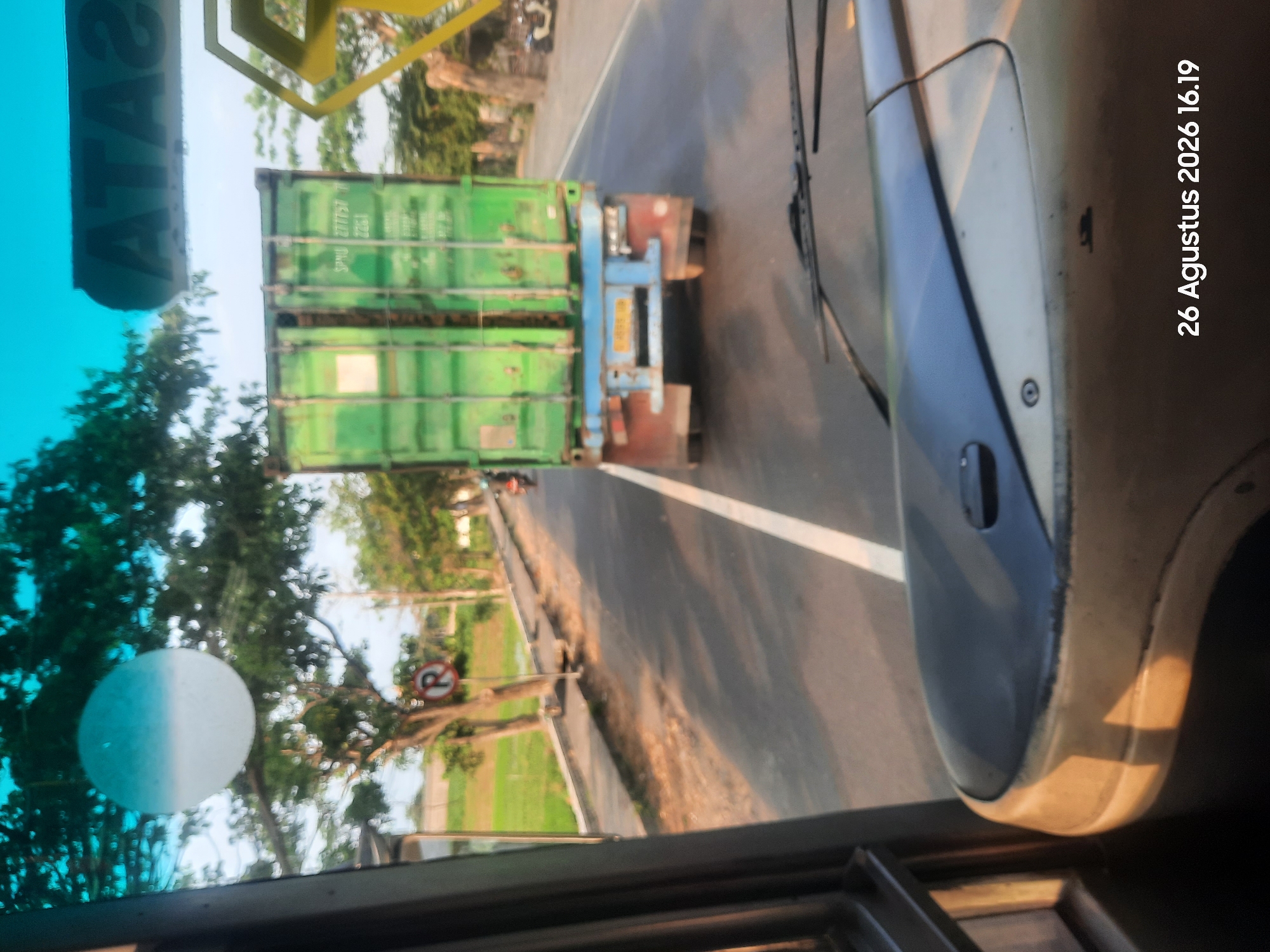 The image shows a view from inside a vehicle, looking out at a road. A green shipping container is being transported on the back of a blue truck. The truck is on the right side of the road, and there is a white line separating lanes. In the background, there are trees and a grassy field. There is also a sign with the letter "R" inside a red circle, indicating no parking. The sky is blue and the sun is shining. The date stamp on the bottom right of the image reads "26 Augustus 2026 16.19". The location is described as Klaseman, Indonesia. The foreground shows a portion of the vehicle's interior, including part of the dashboard and windshield wipers.