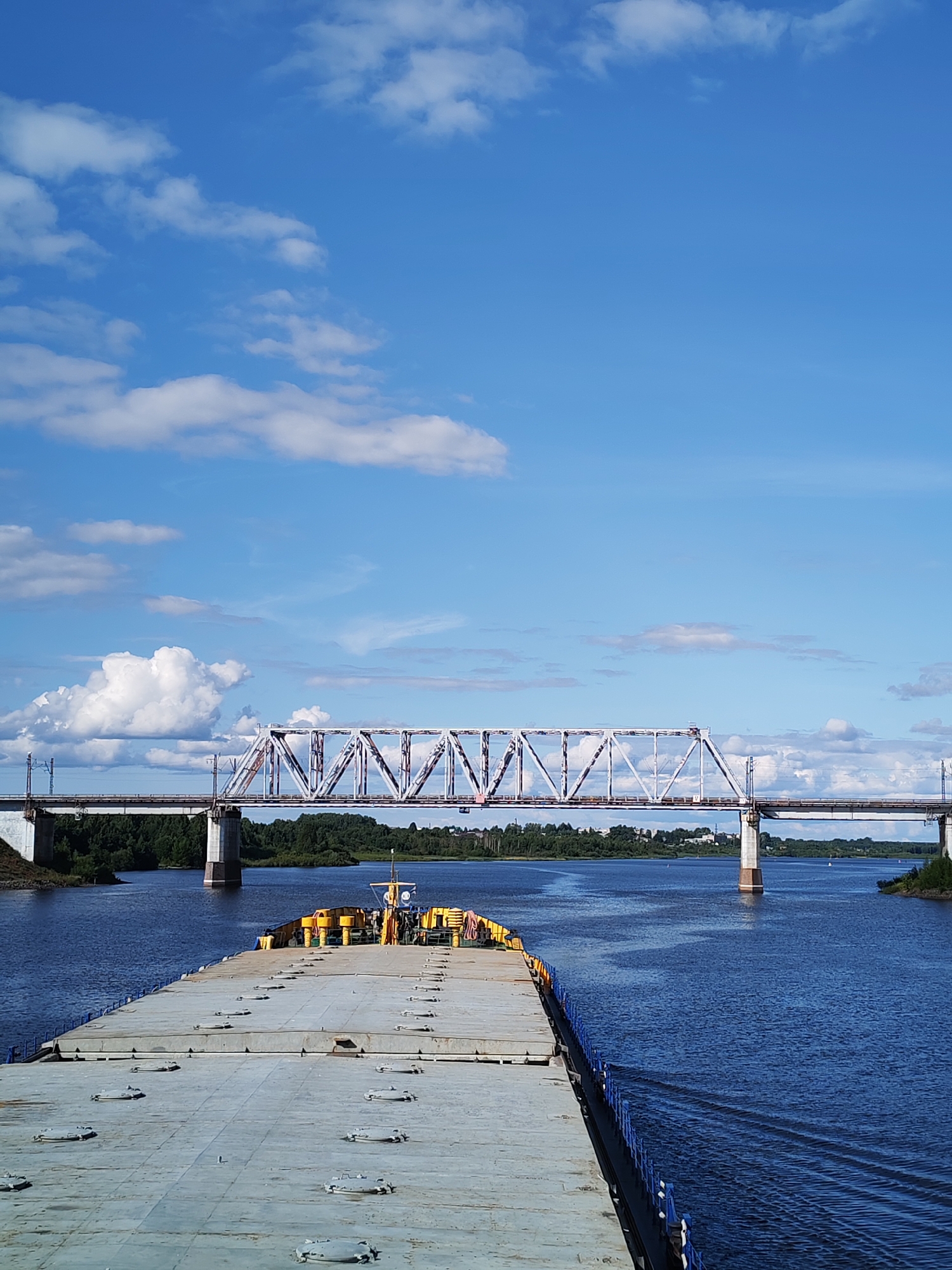 This is a vertical, eye-level, medium shot of a large, flat-bottomed barge moving down a wide, blue river. The barge is the primary subject in the foreground and takes up the lower half of the image. Its deck is gray and appears to be made of metal, with numerous round, recessed hatches spaced evenly across its surface. Along the right edge of the barge, a blue railing with white posts is visible.
In the middle ground, a massive metal truss bridge spans the river. The bridge is painted white and has a complex, geometric structure of interconnected beams. It rests on substantial concrete piers. On the far bank of the river, a dense line of green trees forms the horizon, with hints of buildings and structures peeking through in the distance. The river itself is a deep blue, reflecting the sky.
The sky above is a vibrant blue, dotted with fluffy white clouds, some of which are quite large and billowy, while others are wispy streaks. The lighting suggests it is daytime, likely mid-morning or afternoon, with bright sunshine casting clear shadows. The weather appears to be fair and pleasant. There is no visible text in the image. The scene evokes a sense of journey and transport on a calm waterway.