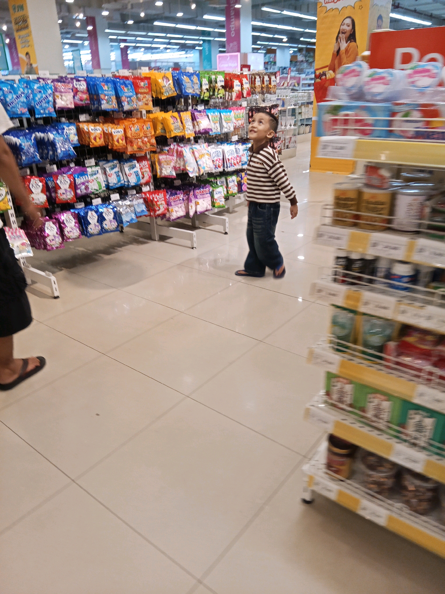 The image shows an indoor scene of a supermarket aisle, likely in Lubuklinggau, Indonesia. A young boy wearing a striped long-sleeved shirt and dark blue jeans is walking away from the camera and looking up towards the shelves. He is on the right side of the frame. To his left, a person's legs, wearing a dark skirt or shorts and flip-flops, are visible, suggesting they are walking in the opposite direction. The shelves are stocked with various packaged goods, including bags of candy or snacks in colorful packaging on the left and various canned and boxed items on the right. The flooring is tiled in a light cream color. The overall impression is a casual shopping trip in a brightly lit retail environment.