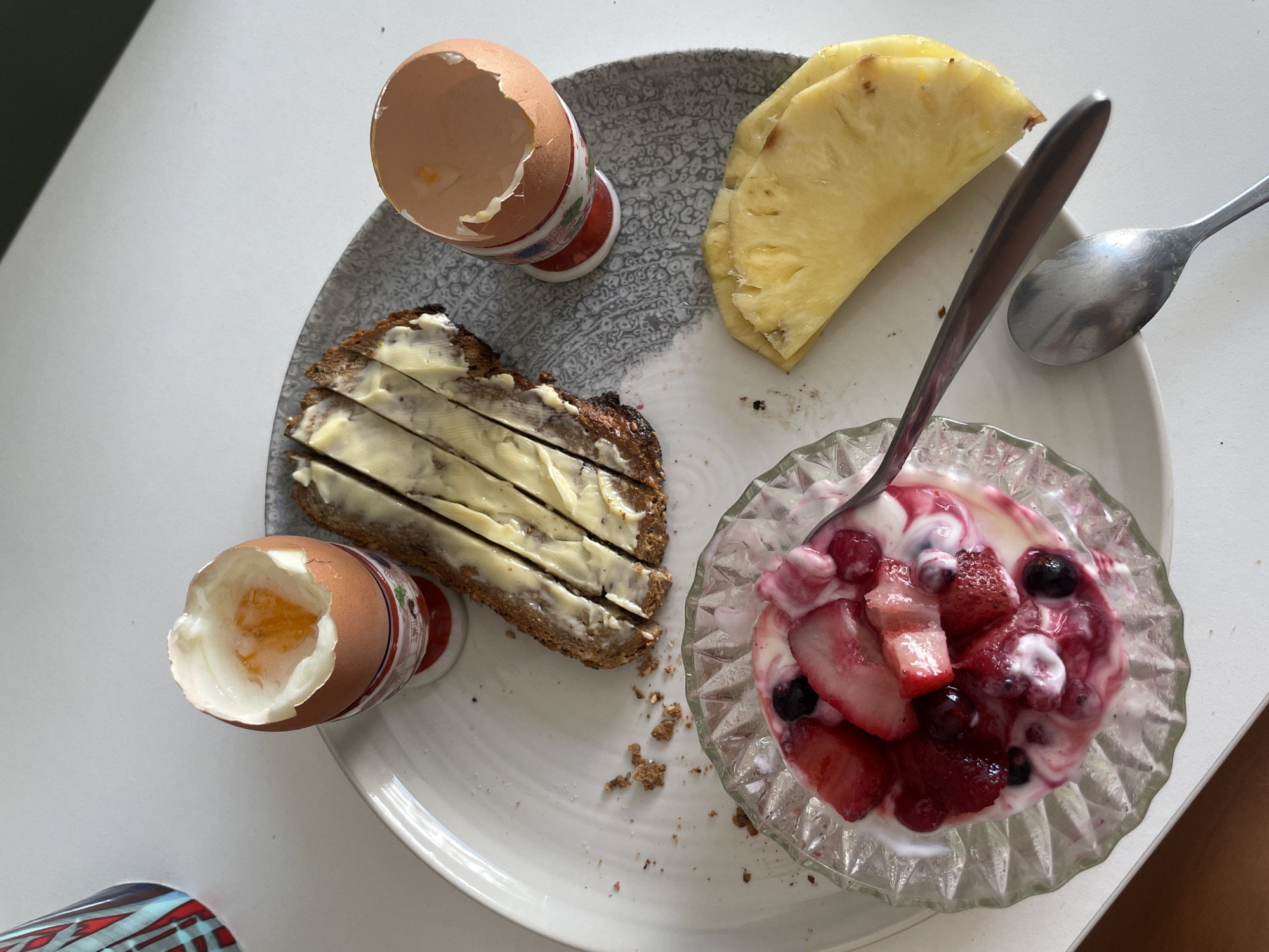 A plate of food is centered on a white surface. The plate contains: two soft-boiled eggs in egg cups, a slice of buttered bread, two slices of pineapple, and a glass bowl of yogurt with strawberries and blueberries. Two spoons are also on the plate. There are no people in the image. The environment is a simple indoor setting. There are no indications the location is Paris, France.