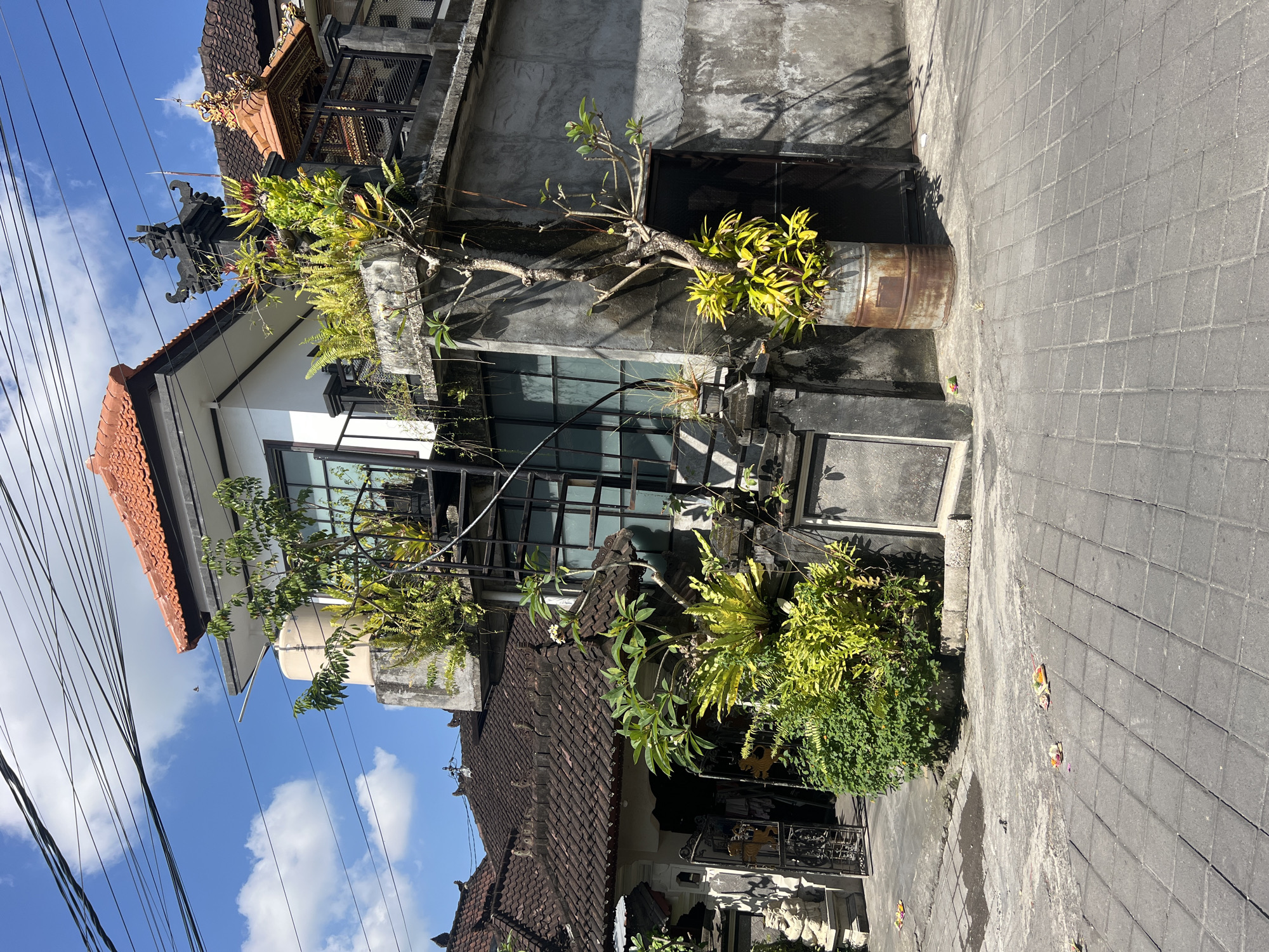 The image captures a street scene in North Kuta, Indonesia, featuring a multi-story building with a mix of traditional and modern architectural elements. The building has a dark-tiled roof on one section and a white section with large, black-framed windows. Decorative carvings are visible near the roofline. A large potted plant in a rusty metal container sits adjacent to the building. A variety of foliage grows around the building. The street is paved with gray brick. Power lines are visible against a blue sky with scattered clouds.