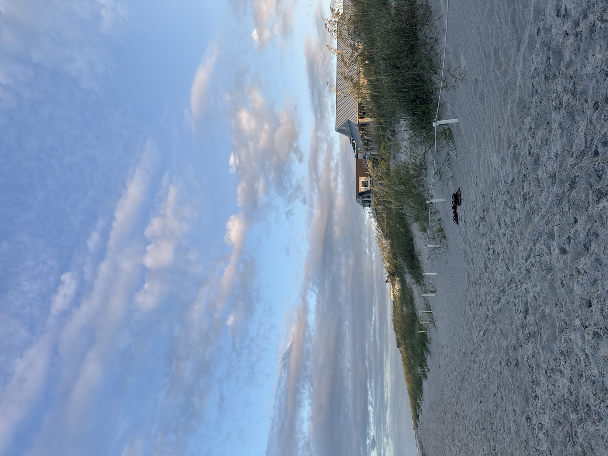 The image depicts a serene beach scene in Jacksonville Beach, United States of America. The foreground is dominated by a sandy beach with gentle tracks and a rope barrier demarcating the edge of the dune line. A pair of flip-flops rests on the sand near the rope. Behind the dune line, tall, wispy beach grass grows abundantly, obscuring much of the view. Several modern houses with metal roofs are visible nestled amongst the vegetation on the elevated dune. The sky above is a blend of clear blue and scattered white and gray clouds, suggesting either early morning or late afternoon. The overall impression is one of tranquility and natural beauty.