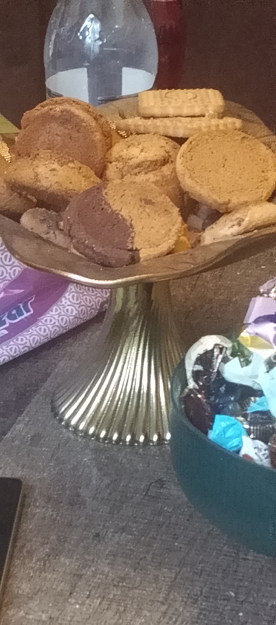 The image depicts a close-up, slightly blurry shot of a golden, fluted cake stand filled with assorted cookies and candies. The cake stand is positioned on a dark, textured surface, likely a table or countertop. In the foreground, to the right, a green bowl overflows with individually wrapped candies, featuring wrappers in shades of blue, purple, and silver. Behind the cake stand, a large, clear glass bottle is visible, containing a clear liquid, possibly water or alcohol, with a white label partially obscured. The background is dark and out of focus. The scene appears to be indoors, possibly a domestic setting or a party. There are no people visible in the image. The lighting is dim, casting shadows and giving the scene a warm, inviting, but somewhat grainy quality. There is no specific information in the image to determine the location within the city of Yerevan, Armenia.