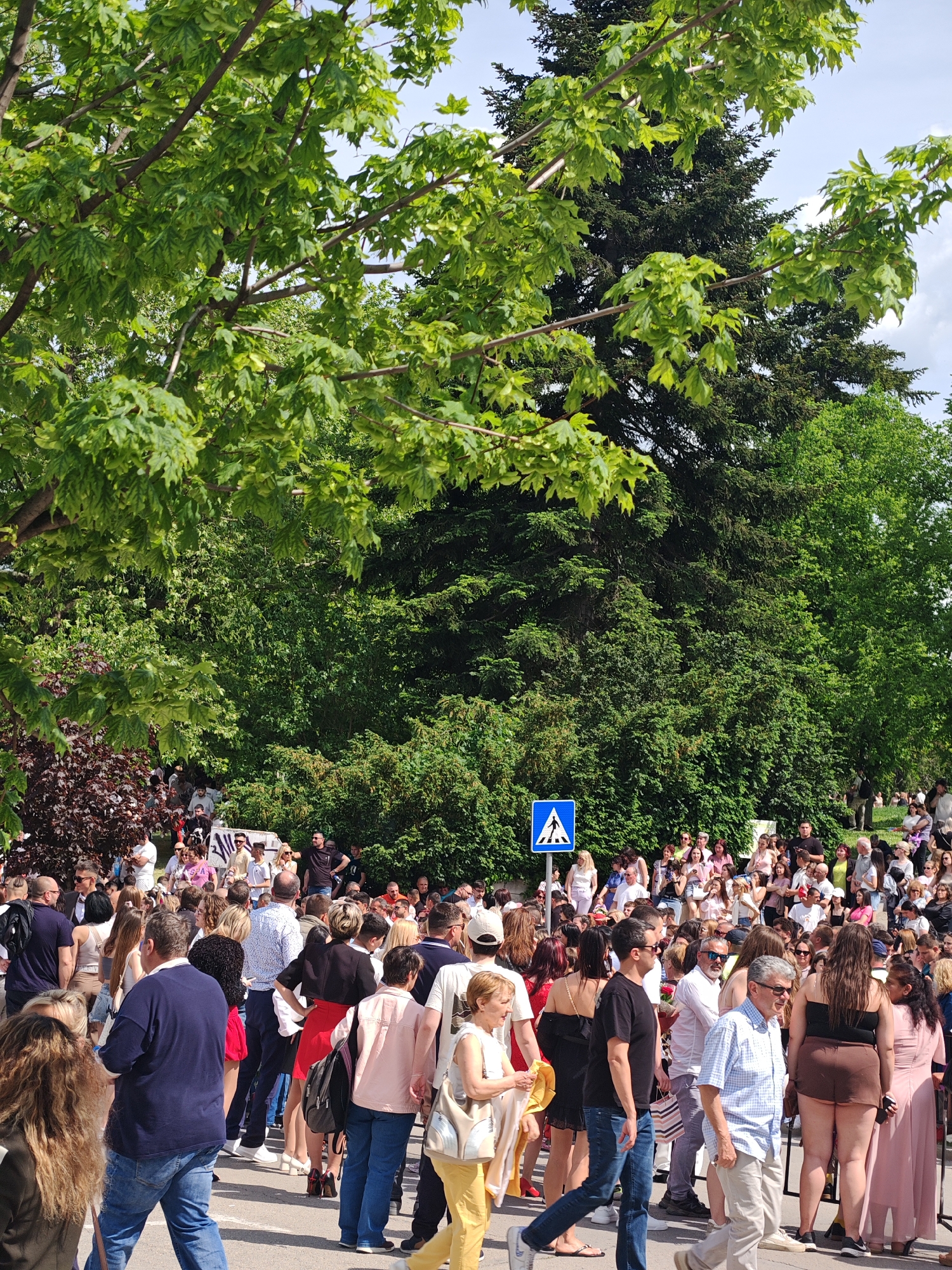 The image shows a large crowd of people gathered outdoors in Shumen, Bulgaria. The crowd is a mix of ages and genders, dressed in casual attire. A blue pedestrian crossing sign is visible amongst the crowd. Lush green trees and foliage surround the crowd. The people are standing and walking, some interacting with each other. The scene appears to be a public gathering on a sunny day.