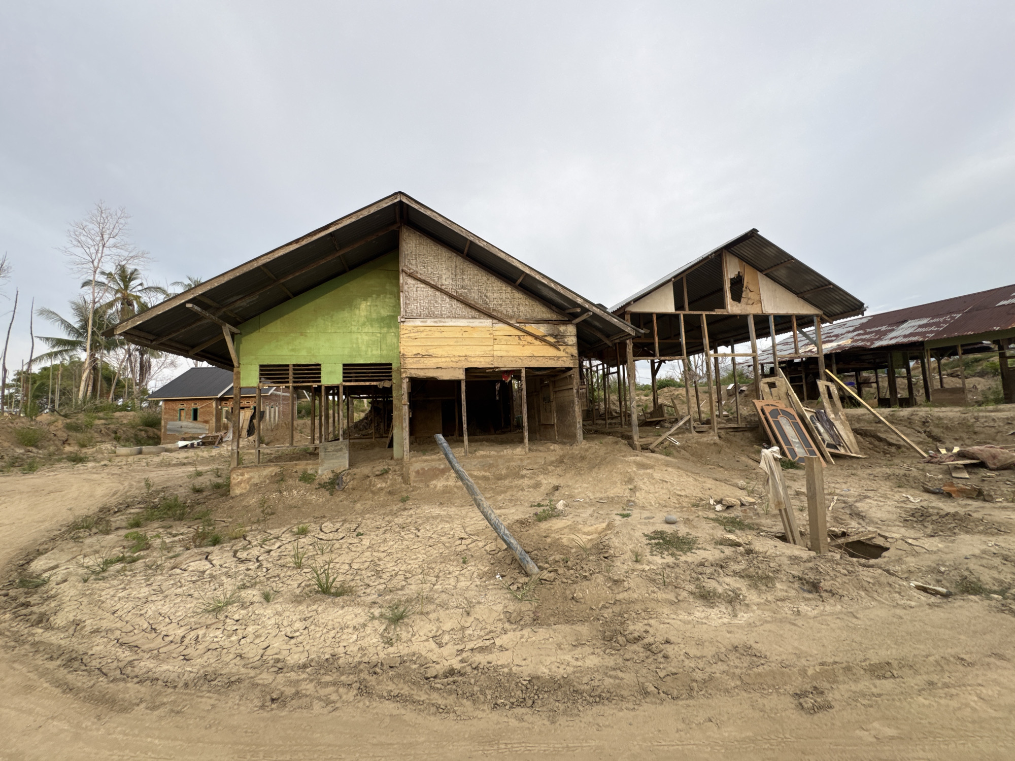 The image depicts a rural or semi-rural scene in Meunasah Raya, Indonesia, characterized by a pair of partially constructed or dilapidated wooden structures. The foreground is dominated by dry, cracked earth with sparse vegetation, suggesting a possibly arid or drought-affected environment. A dirt path or road is visible in the lower portion of the frame.
The structures themselves are elevated on stilts and feature corrugated metal roofs. The first structure on the left is partially painted in green and yellow, with exposed wooden framing and what appears to be woven bamboo paneling. The second structure to the right is more open, revealing its skeletal wooden framework and a damaged interior wall. Further back, another structure with a rusty corrugated metal roof is visible.
Scattered around the site are various construction materials and debris, including wooden planks, what seem to be doors leaning against a structure, and other miscellaneous items. A tilted metal pipe or pole is embedded in the dry earth in front of the main structures. The overall impression is one of ongoing construction, abandonment, or post-disaster recovery. The sky is overcast, contributing to a muted lighting condition. No people are visible in the image.