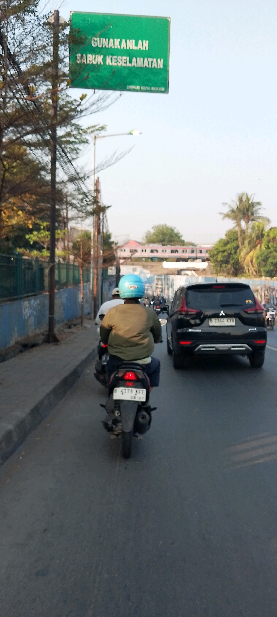 In Bekasi, Indonesia, a motorcycle rider wearing a light brown jacket and a blue helmet navigates a street. The rider is on a black motorcycle with a license plate reading "B 4378 KFE". Behind the motorcycle, a black SUV with license plate "B 2344 KYB" is visible. A green sign with white text reading "GUNAKANLAH SABUK KESELAMATAN" (which translates to "USE SEATBELTS") is mounted on a pole to the left of the frame, with smaller text below indicating "DINHUB KOTA BEKASI" (Department of Transportation, Bekasi City). The environment suggests an urban setting with trees lining the street and buildings in the background. The sky is clear, indicating daylight.