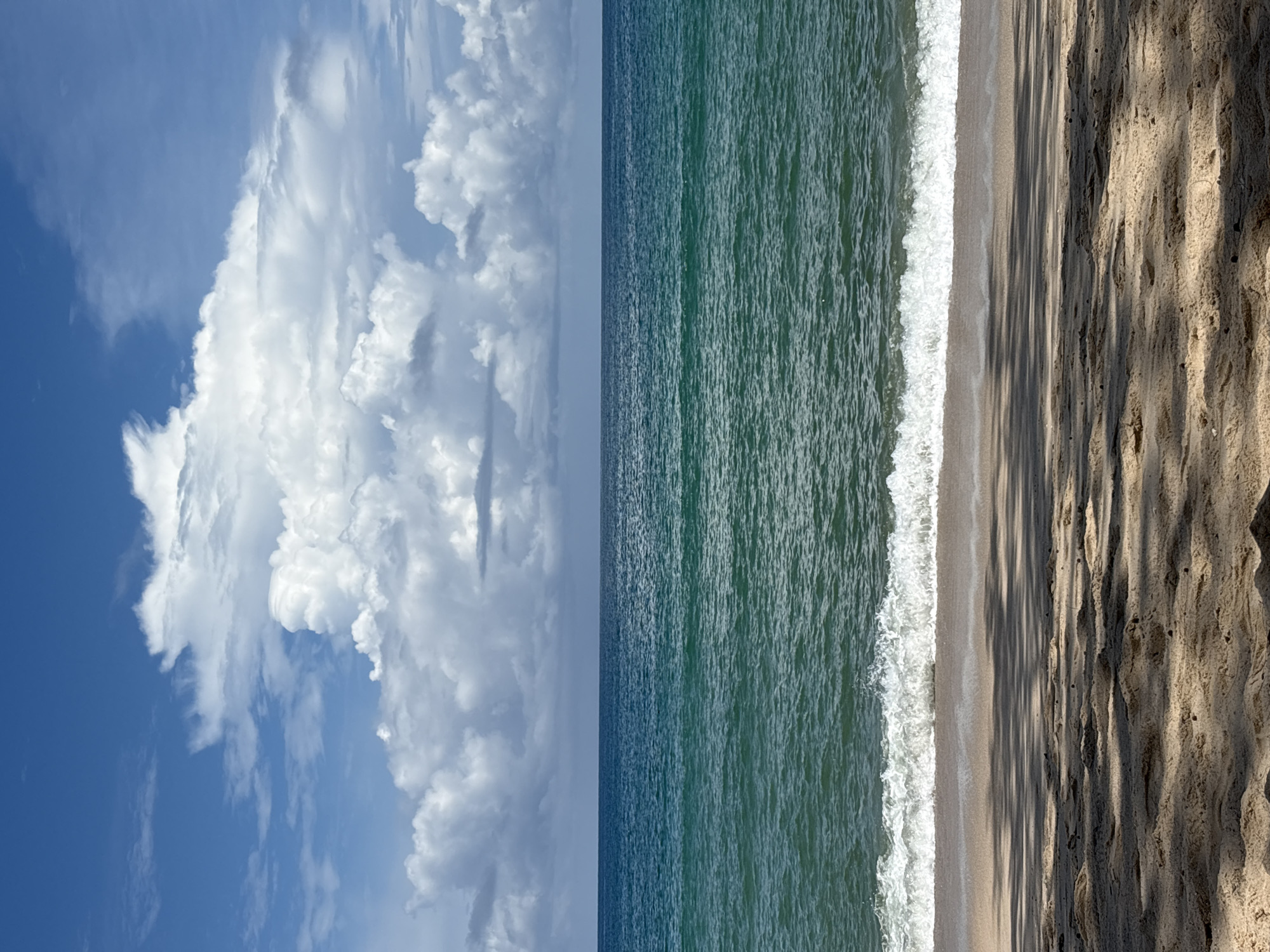 A wide shot of a beach in Choeng Thale, Thailand, shows a sandy shoreline in the foreground, with dappled shadows cast by unseen trees. The ocean stretches out in the middle ground, a calm expanse of turquoise water with a gentle wave breaking near the shore. In the background, a clear blue sky is dotted with large, fluffy white clouds. The horizon line is sharp, separating the sea from the sky. There are no people or man-made objects visible in the frame.