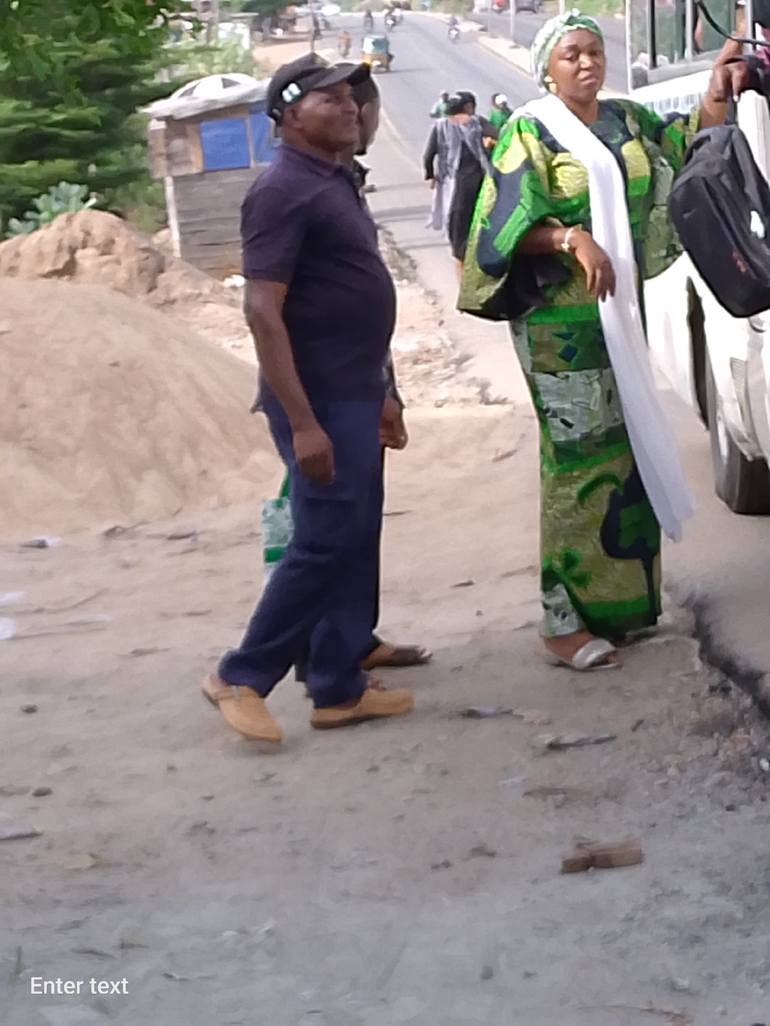 The image depicts a street scene in Adankolo, Nigeria. A man in a dark blue polo shirt, dark blue pants, a dark baseball cap, and tan-colored shoes is walking along a dusty street. Beside him stands a woman wearing a green and black patterned dress, a white scarf, and a matching headwrap. She is holding a black bag and appears to be interacting with a vehicle, possibly boarding or disembarking. A pile of sand is located to the left of the man, and a small structure with blue trim stands further back. In the distance, vehicular traffic, including motorcycles and a three-wheeled vehicle, can be seen on a road. Additional individuals are also visible in the background.