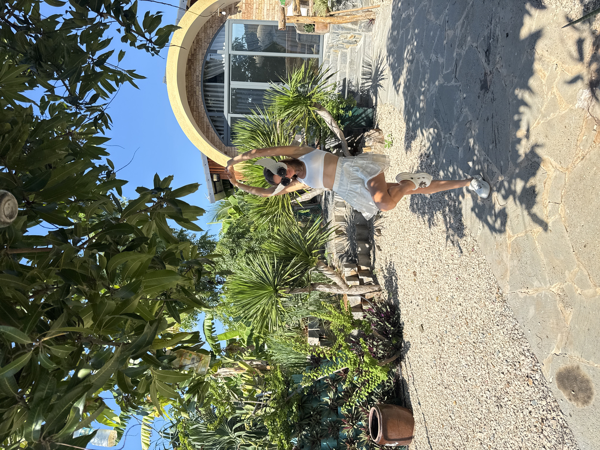 In Mũi Né, Vietnam, a woman stands on a stone pathway in a courtyard with a lush garden setting. She is wearing a white crop top, a short white skirt, and white platform shoes. Her arms are raised above her head, and she is balancing on one leg. She is also wearing large, decorative sunglasses with white wing-like accents. The courtyard is covered in light-colored gravel and features a large, brown ceramic pot on the left. In the background, a building with a distinctive arched entrance and large glass doors is visible. The garden is filled with various tropical plants, including palm trees and other dense foliage. The sky is clear blue, indicating a sunny day.