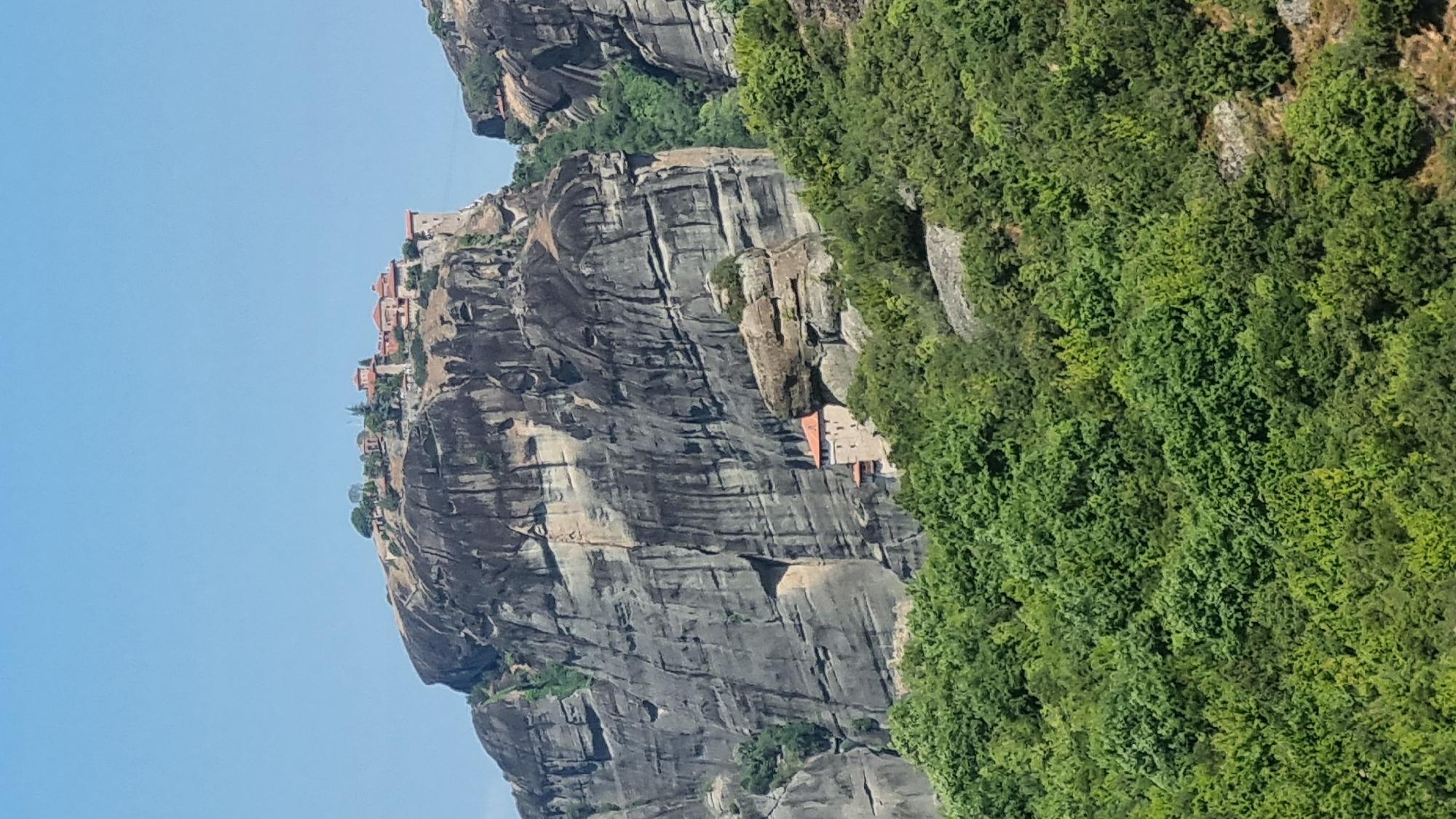 The image shows the Meteora Monasteries, a UNESCO World Heritage site, located near Kalambaka, Greece. The scene features towering sandstone rock formations, often called "pillars" or "mountains," with several monasteries perched precariously on their summits. The rock faces are vertical and rugged, showing signs of erosion. Lush green vegetation covers the lower slopes and the terrain surrounding the rock formations.
In the upper portion of the image, a monastery complex with red-tiled roofs is visible on the flat summit of a large rock. This monastery appears to be a collection of buildings, possibly interconnected.
In the middle ground, on the side of a rock formation, a smaller stone building with a red roof is visible. This structure is built directly into the rock face, seemingly attached to it.
The sky is clear and blue, indicating a sunny day. The overall impression is one of awe-inspiring natural beauty combined with human ingenuity and religious devotion, showcasing the unique architecture of the Meteora monasteries against a dramatic geological backdrop. There are no visible people or animals in the frame.