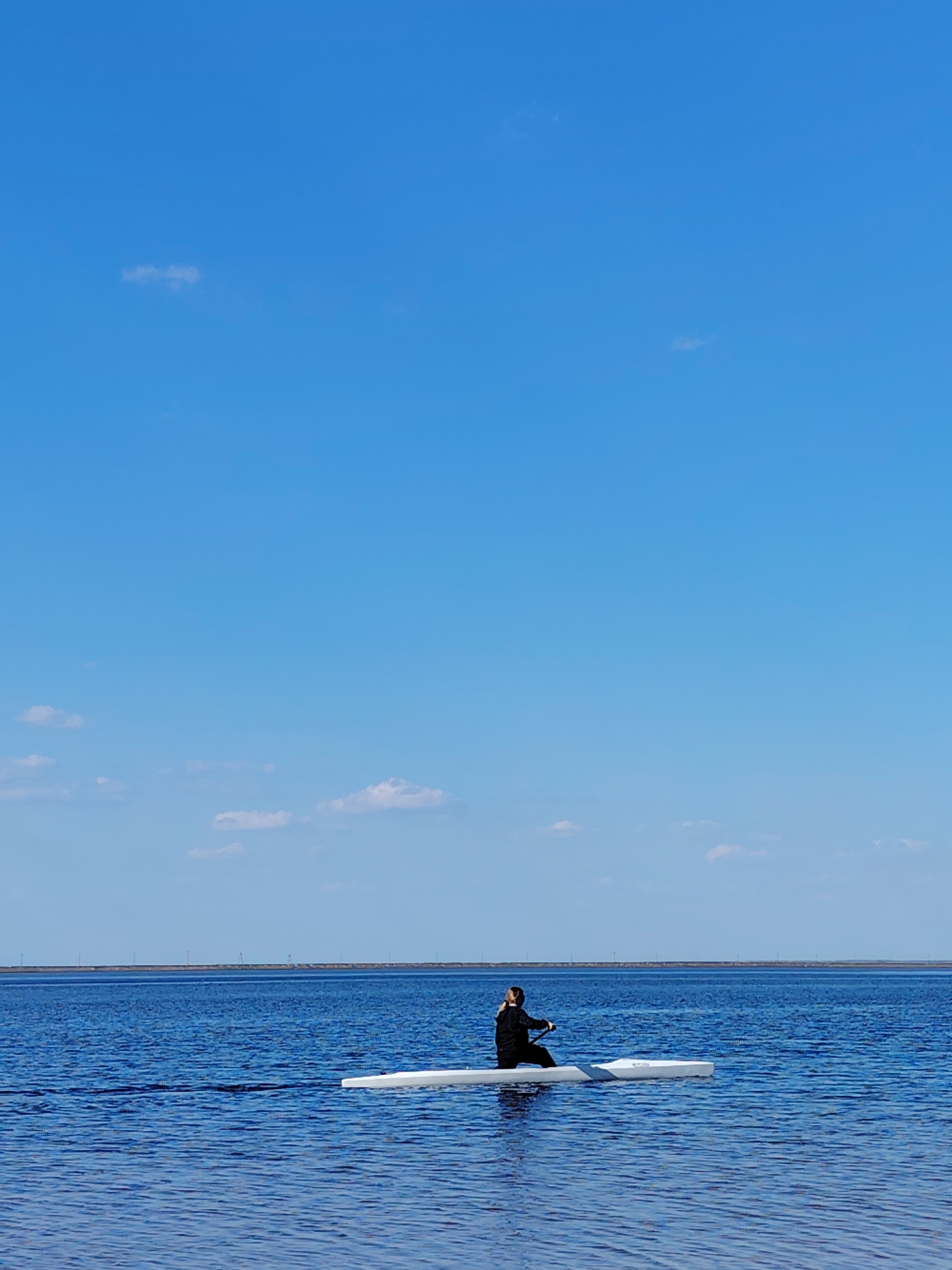A long, narrow white kayak is on blue water under a clear blue sky with scattered clouds. A person is seated in the kayak, wearing dark clothing, possibly rowing or paddling. In the distance, a visible landmass can be seen along the horizon. The image was taken in the city of Chaikovsky, Russia.
