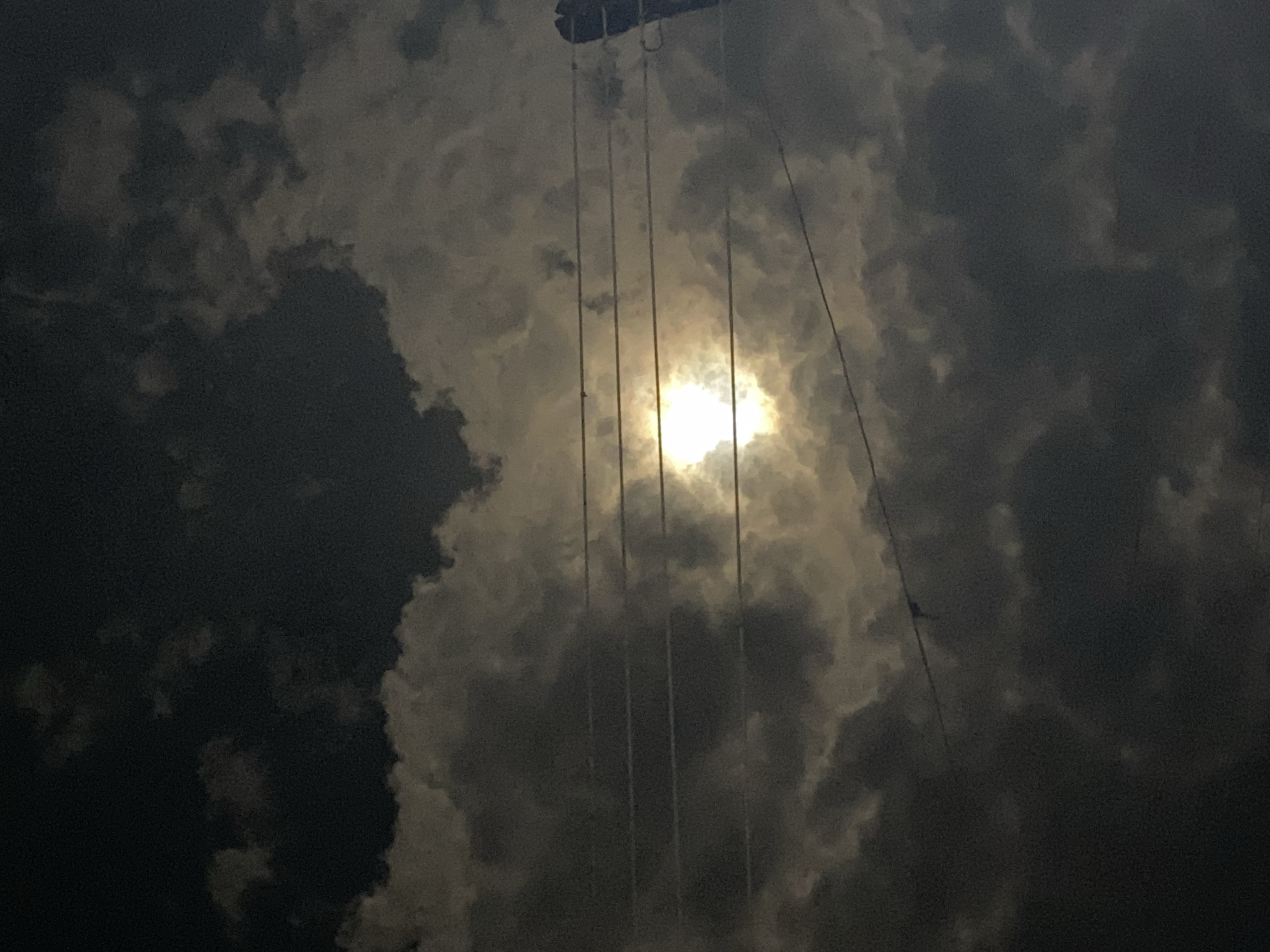 The image shows a night sky in Maigizo, Nigeria. The scene is dominated by dark, heavy clouds, partially obscuring a bright light source, likely the moon. Wires or ropes are visible stretching vertically across the frame, partially obscuring the light source and suggesting the presence of overhead infrastructure. The overall environment is dark and atmospheric.