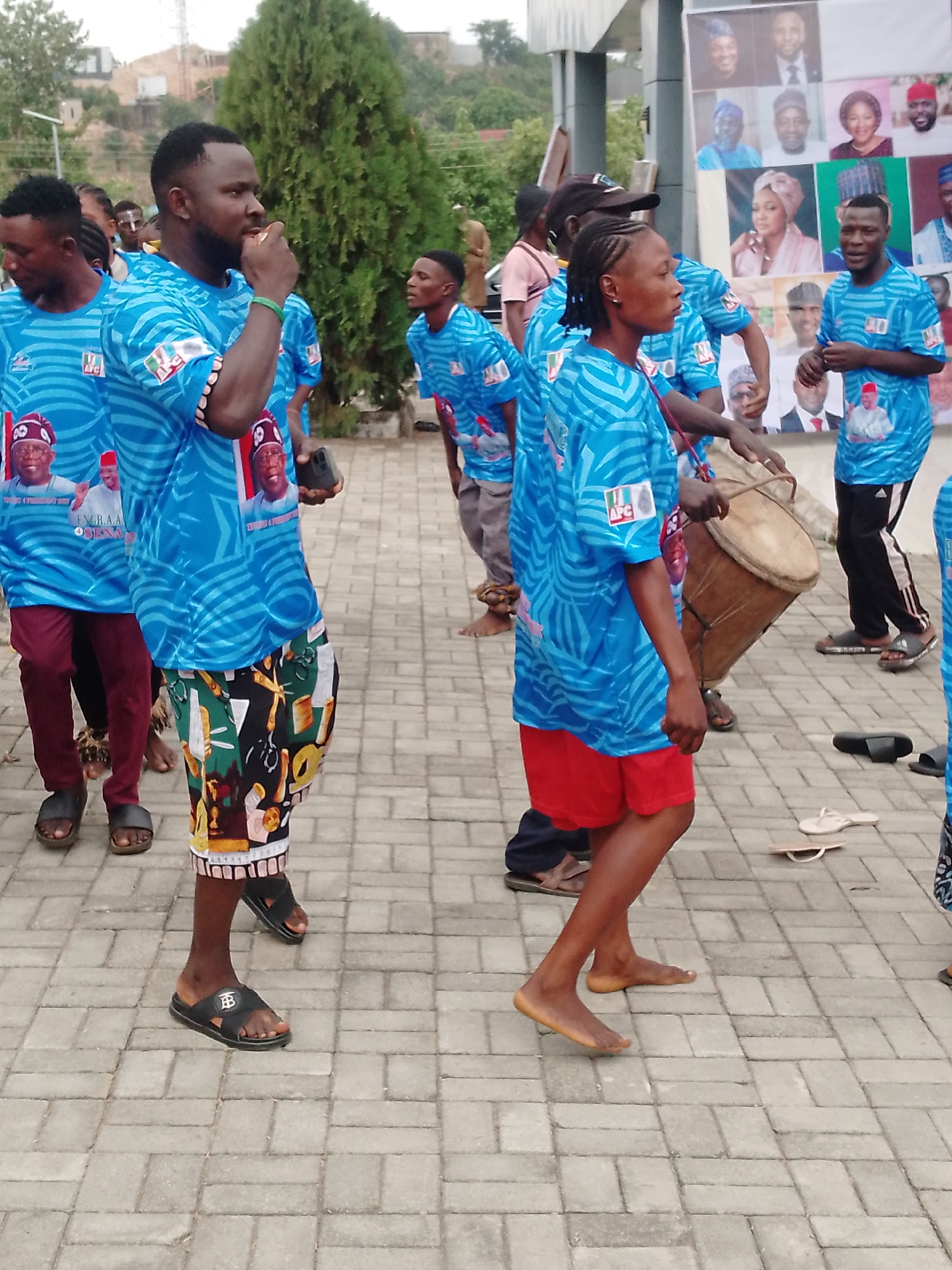 A street in the city of Unknown, Unknown, is filled with people wearing similar light blue t-shirts adorned with a portrait of a man. Some are also wearing colorful shorts or dark trousers. One individual is playing a drum, while others are clapping or looking around. The ground is paved with rectangular tiles. A tree and a building are in the background, and a poster with portraits of multiple people is visible on the building.
