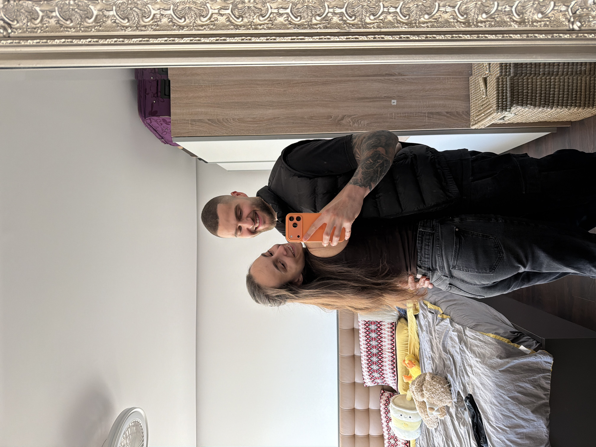 This full-length mirror selfie captures a smiling couple in what appears to be a bedroom in Sofia, Bulgaria. The image is taken from the perspective of their reflection in a large mirror with an ornate, possibly silver or gold-colored, frame at the top.
The man, on the left, has a shaved head, a beard, and is wearing a black t-shirt and a black puffy vest. His left arm, visible with tattoos, is extended, holding an orange smartphone to capture the photo. He is smiling broadly, looking towards the camera (his reflection).
Leaning into him from the right is a woman with long, brown hair. She is wearing a dark top and dark jeans, and is also smiling, gazing towards their reflection.
The background, visible through the mirror, reveals elements of a cozy room. Behind the couple, there's a bed with a beige upholstered headboard. The bed is covered with a grey duvet, a yellow pillow, and pillows featuring a red, white, and black geometric pattern. Several stuffed animals, including a brown bear, a yellow duck-like toy, and a small white and yellow plushie, are arranged on the bed. To the far left, a white standing fan is visible. Further in the background, a wooden wardrobe or drawer unit, a purple suitcase or bag, and a woven wicker basket are also reflected.
The room is brightly lit, suggesting it is daytime. Both individuals appear happy and relaxed, enjoying the moment as they capture their reflection. No visible text is discernible in the image.