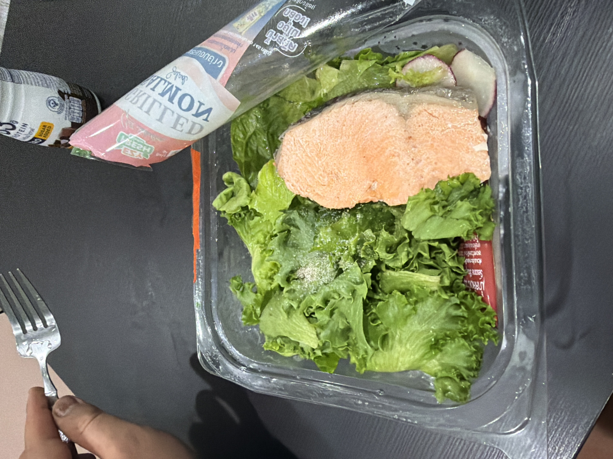 A person's hand holds a fork over a clear plastic container filled with a salad. The salad consists of green lettuce, a piece of cooked salmon, and sliced radishes. A pink and white tube, likely a sauce or dressing, rests on top of the salad and is partially covering it. In the background, a white bottle with text is visible. The scene appears to be set on a dark, textured surface, possibly a table. The image was taken in Baan Mae Nam, Thailand, suggesting a meal being prepared or consumed in that location.