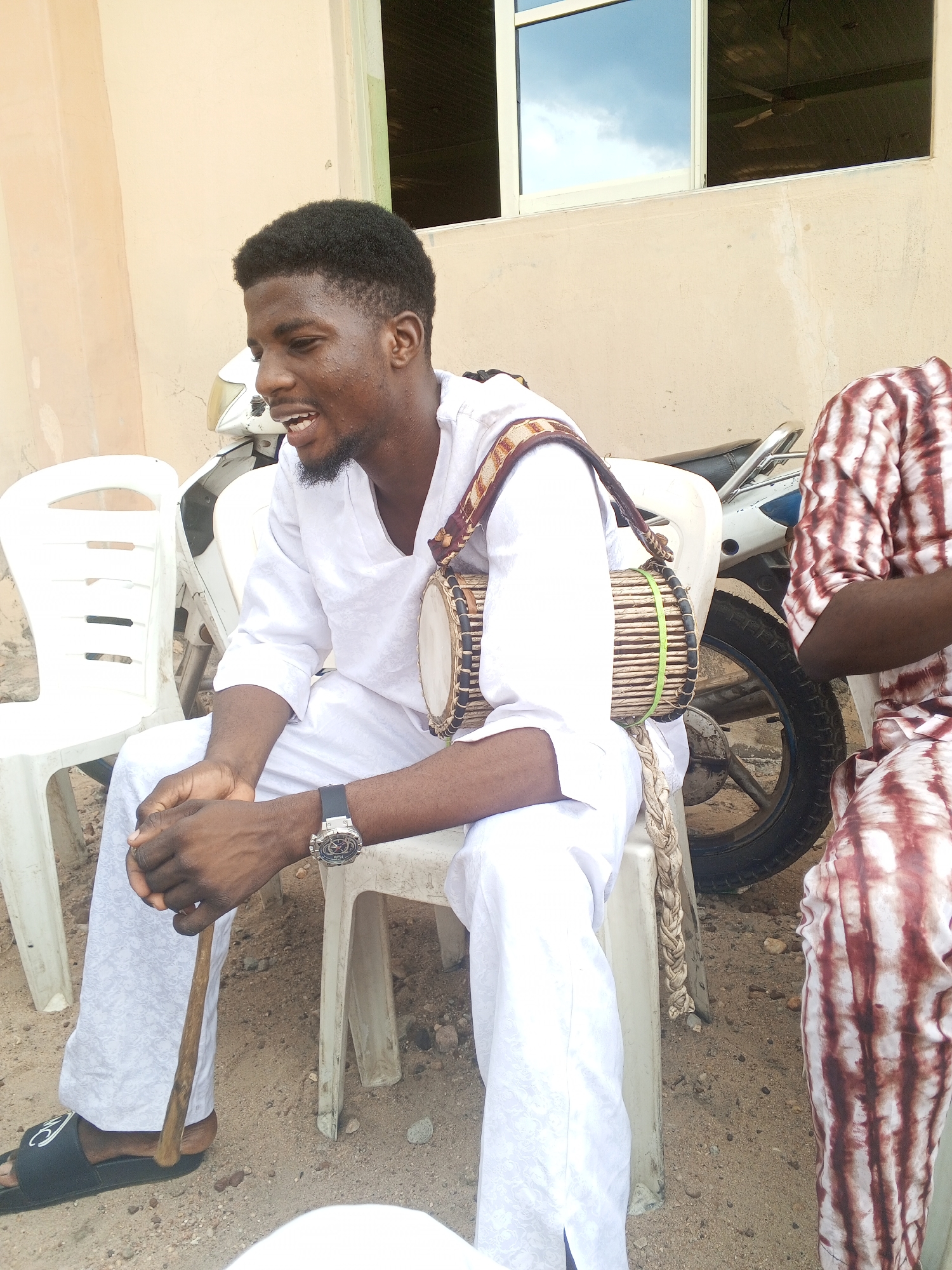 A man sits on a white plastic chair outdoors in Zango-ndaji, Nigeria. He is wearing a white shirt and pants, black sandals, and a watch. A small drum with a woven strap is slung over his shoulder. He holds a wooden stick in his hands. Behind him is a white motorcycle. A portion of another person wearing a brown and white patterned shirt is visible on the right. In the background is a wall with a window. The ground appears to be dirt.