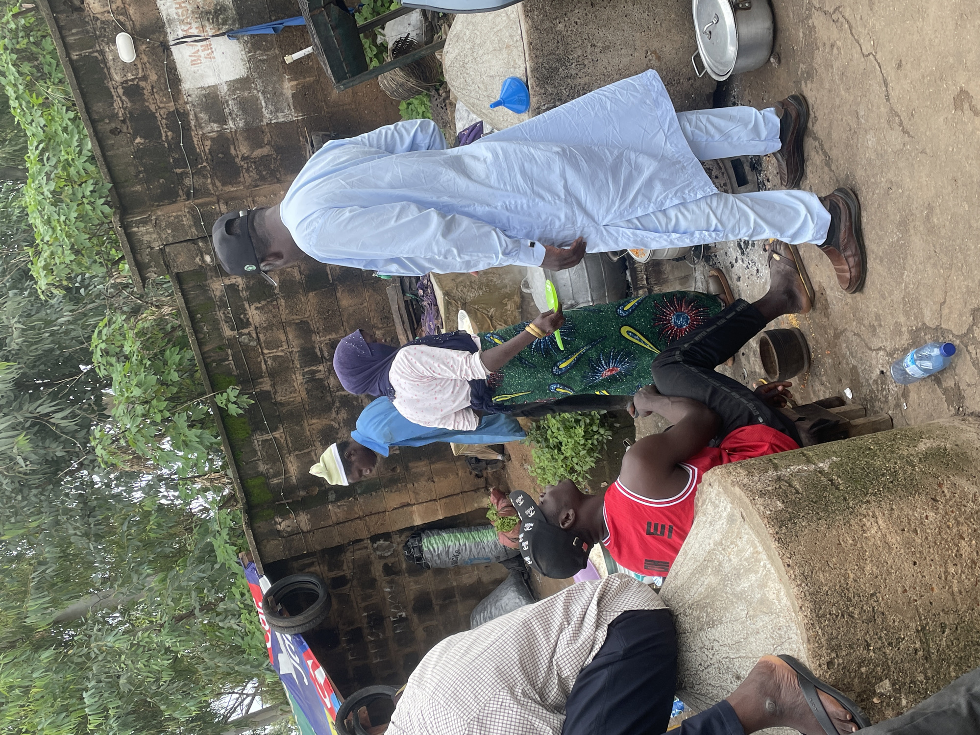 A medium shot captures a group of people gathered in what appears to be an outdoor market or food preparation area in Jos, Nigeria. The scene is cluttered with various objects, suggesting a bustling, informal setting. A person wearing a light blue kaftan and a black baseball cap stands facing away from the camera, engaged in conversation with another individual. To the left, a person in a red tank top and black pants is seated on a large, rounded concrete structure, looking upwards. Another individual, partially visible, is seated to the far left, wearing a checkered shirt and black pants. A woman in a green patterned skirt and a headscarf is also present, holding a green ladle. In the background, a brick wall with some graffiti and a light fixture is visible, along with dense foliage. Several large pots and cooking vessels are scattered around, and a blue plastic funnel is placed on a concrete mound. A plastic water bottle lies on the ground in the foreground. The ground surface is rough and uneven, indicative of a dirt or paved area common in such environments. The lighting suggests an overcast or shaded day.