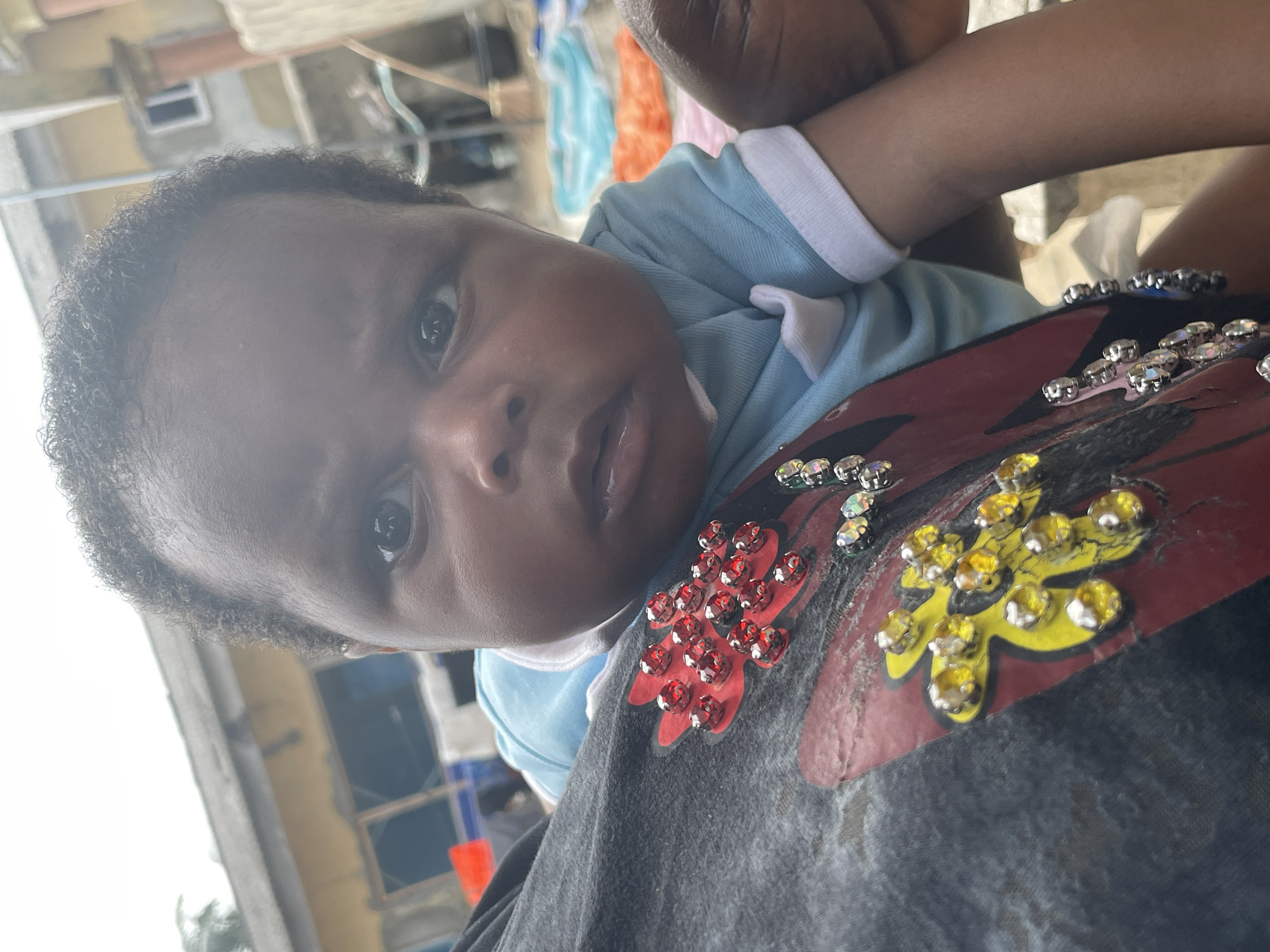 A close-up, slightly tilted portrait shows a baby being held by an adult, likely in an outdoor setting in Badagry, Nigeria, during the daytime.
The primary subject is a baby with dark, curly hair and dark, observant eyes, looking directly towards the camera with a curious expression. The baby's mouth is slightly open, and their skin tone is dark. They are wearing a light blue top with white cuffs and collar.
An adult's arm and shoulder are visible in the foreground, indicating they are holding the baby. The adult is wearing a dark-colored t-shirt, possibly black or dark grey, which is adorned with decorative embellishments. These decorations consist of clusters of jewel-like stones in red, yellow, and clear/iridescent colors, forming patterns that resemble flowers or leaves on the fabric.
The background is out of focus, revealing hints of structures and what appears to be clothes hanging, suggesting a residential or semi-urban outdoor environment. The bright, diffused lighting indicates it is daytime. No visible text is present in the image. The scene captures a tender, everyday moment between the baby and the unseen adult.