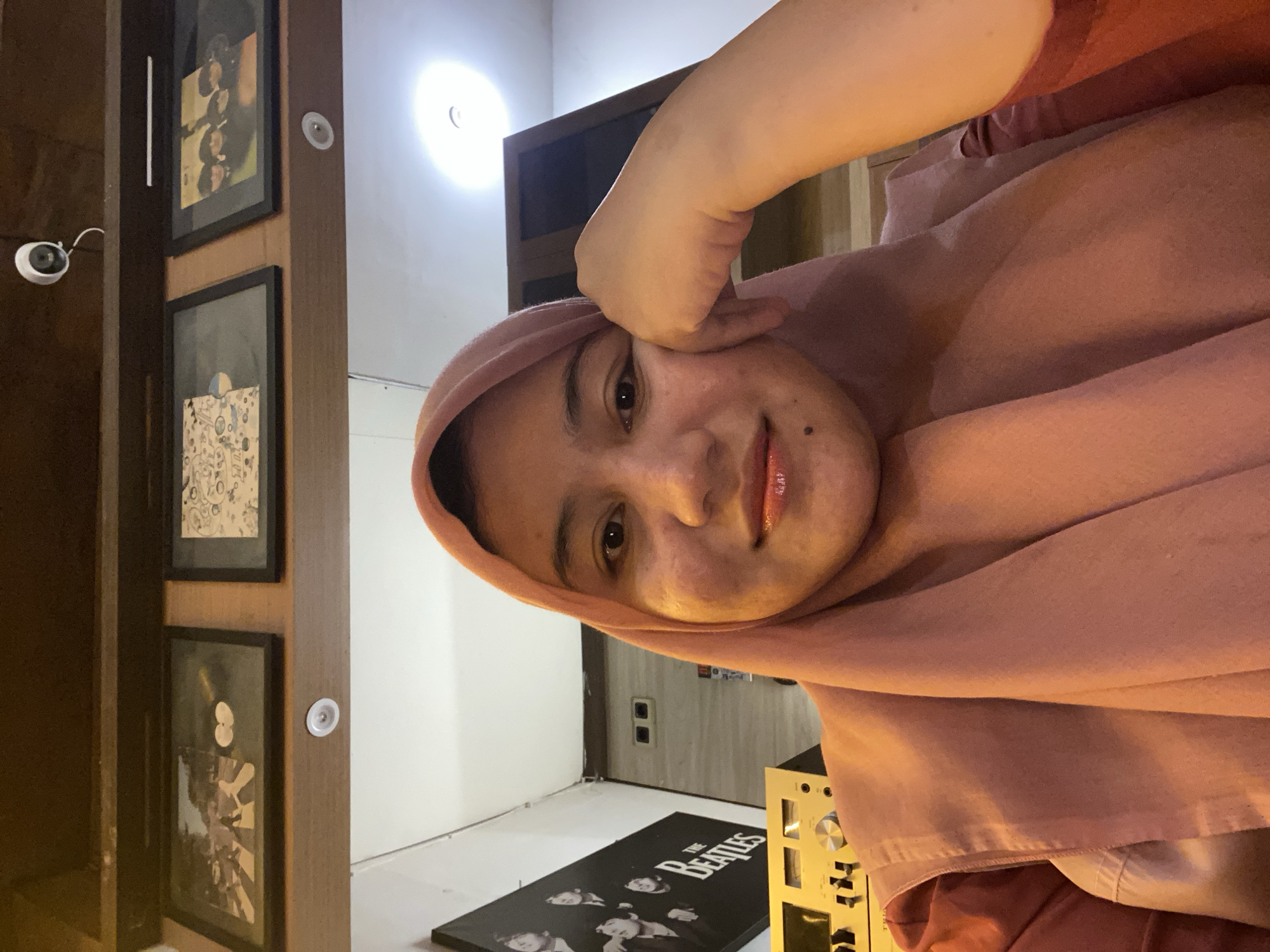 A young woman wearing a pink hijab and a reddish-orange top poses for a selfie in what appears to be a cafe or lounge. The woman is resting her head on her hand, with her eyes looking directly at the camera. Behind her, a black and white poster of The Beatles is visible on the wall, along with framed album covers. The lighting is warm and ambient, suggesting an indoor setting. There is no identifiable information about the location within Banjarbaru, Indonesia.