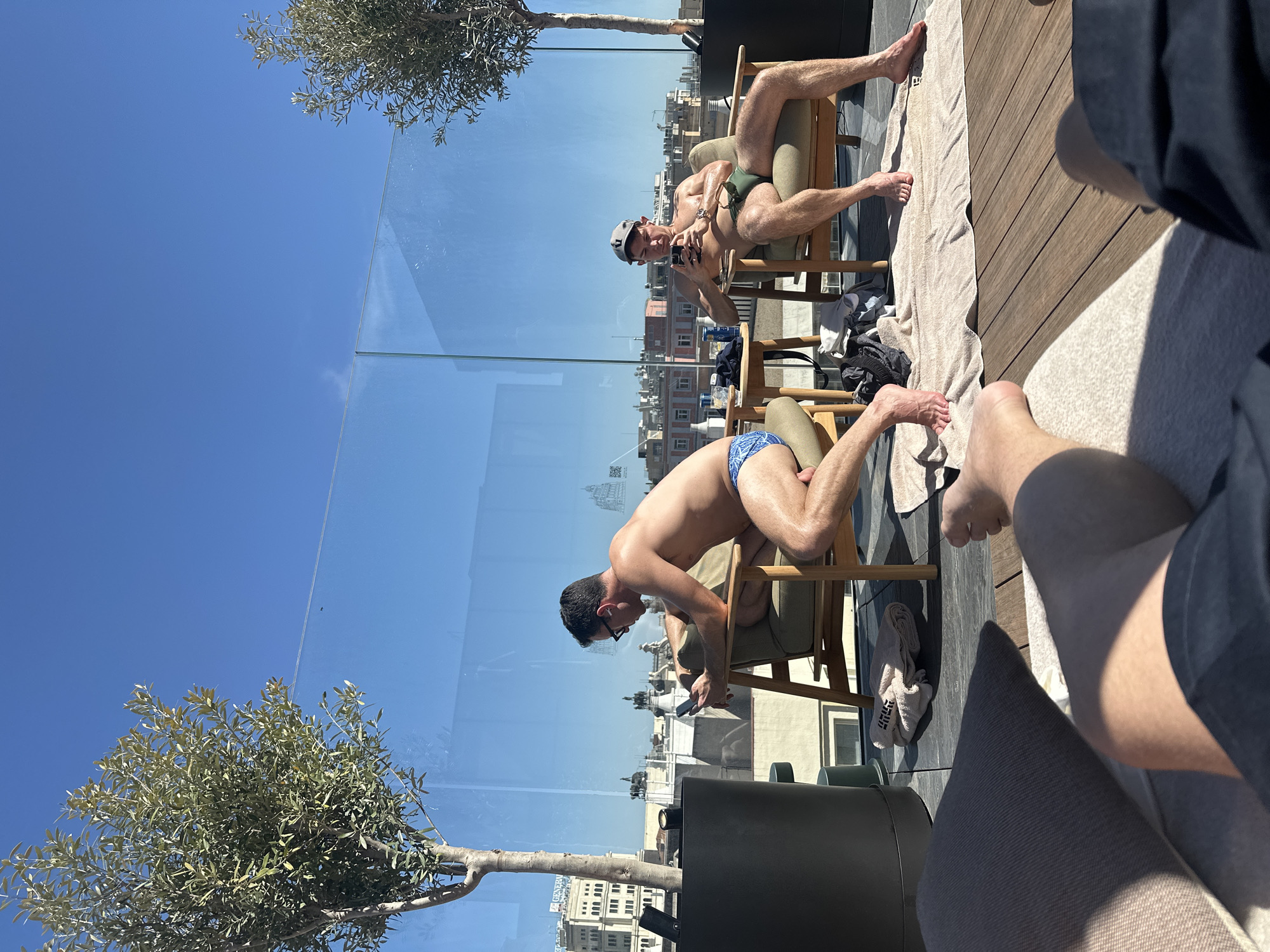The image depicts a bright, sunny scene on a rooftop terrace in Madrid, Spain, taken from a low, first-person perspective. In the immediate foreground, the legs of a person are visible, resting on a cushion.
Two men in swimwear are seated on wooden chairs on the wooden-decked terrace. The man closer to the viewer, wearing blue and white patterned swim briefs and sunglasses, is leaning forward, looking at his phone. Behind him, another man, dressed in olive green swim briefs and a baseball cap, is also engaged with his phone. Towels are laid out on the deck, and bottles are visible on a small table positioned between their chairs.
The terrace features decorative olive trees and is edged by a clear glass balustrade. Beyond and reflected in the glass, a panoramic cityscape of Madrid is visible, including buildings and what appear to be statues and ornate architectural details. The sky above is a vibrant, cloudless blue, indicating a clear, sunny day. Bright sunlight casts strong shadows, suggesting it is midday or early afternoon.
A white towel on the deck has the text "GENERAL" visible, and a QR code is faintly reflected on the glass barrier. The overall atmosphere is one of leisure and relaxation on a warm, summer-like day.