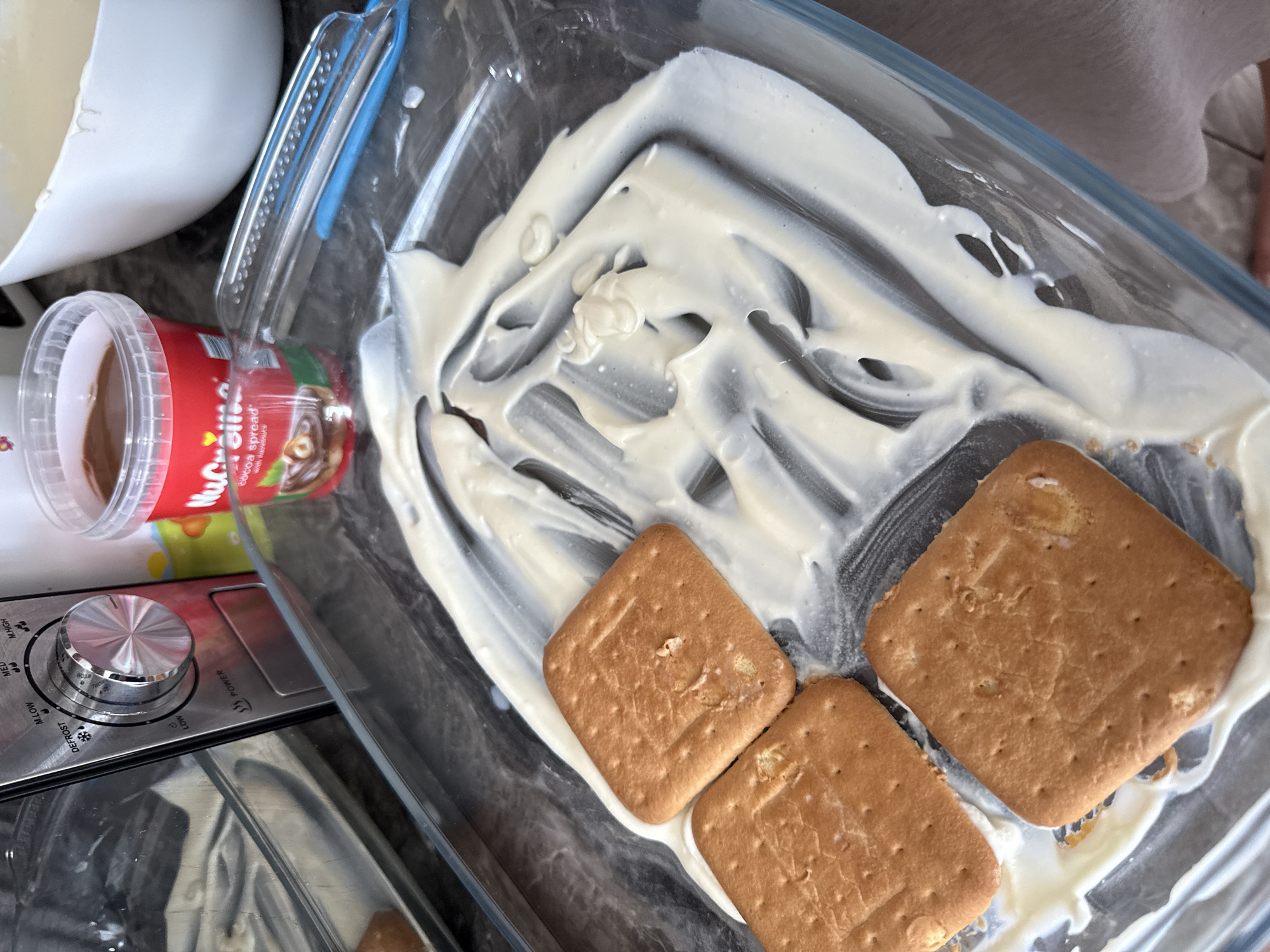 An overhead, close-up shot captures a kitchen scene in Rogosh, Bulgaria, focusing on the preparation of a layered dessert. The central subject is a clear rectangular glass baking dish. Inside, a thick, white creamy substance, possibly yogurt or cream cheese, is spread unevenly across the bottom, showing swirls and peaks. Three light brown, square-shaped biscuits are placed on top of or partially embedded in this creamy layer. The biscuits have a perforated texture.
To the left of the dish, a red plastic container with a clear lid is visible. It contains a dark brown spread, identified by its partially visible label as "cocoa spread" and "with hazelnuts," likely from a brand starting with "Nuc...". A white ceramic bowl is partially seen in the upper left corner. Below the red container, part of a metallic kitchen appliance is visible, featuring a prominent silver control knob. The knob has various power settings marked around it, including "M.HIGH," "HIGH," "MED," "M.LOW," "DEFROST," and "LOW," along with "POWER." The overall lighting suggests an indoor setting, possibly during the daytime. The scene conveys a moment of active food preparation.