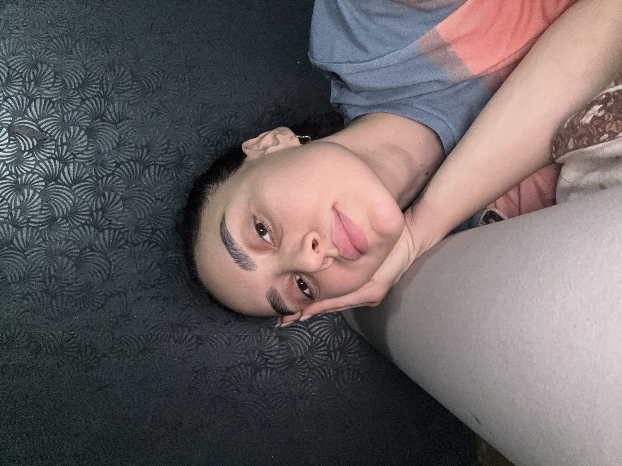 A woman is lying down on a black patterned surface, propping her head up with her hand. She is wearing a shirt with blue, pink, and gray fabric. The location in Tbilisi, Georgia, is not discernible from this image.