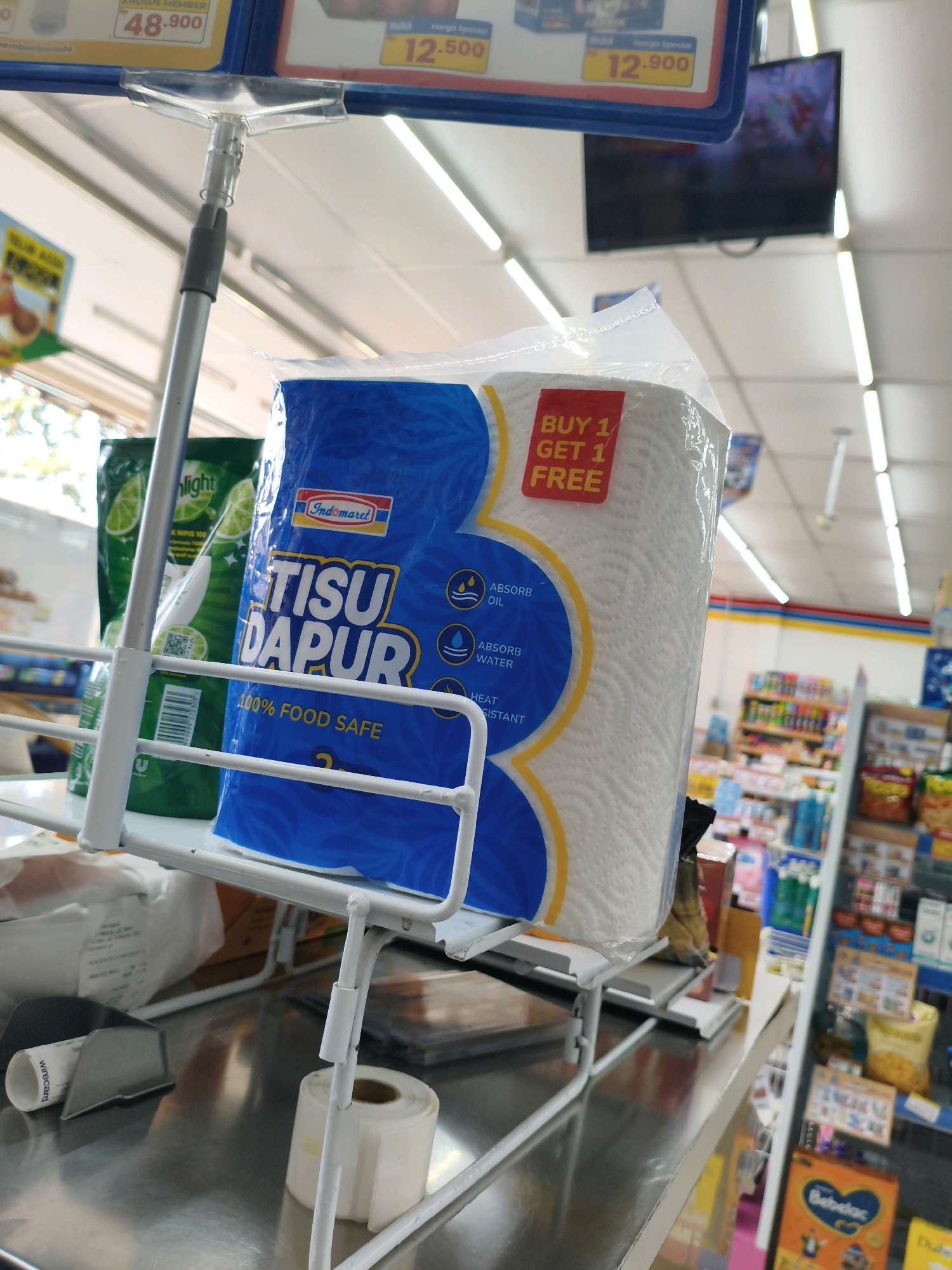 In the city of Sumberejo Kulon, Indonesia, a close-up shot captures a display of paper towels in what appears to be a convenience store. The dominant item is a large roll of white paper towels encased in a clear plastic bag with a blue and yellow label. The label prominently features the text "Indomaret" and "TISU DAPUR", indicating it's a kitchen towel from the Indomaret brand. A red sticker on the packaging reads "BUY 1 GET 1 FREE". To the left of the paper towels, a green package, likely dish soap, is visible. The items are placed on a white metal display rack, which is positioned on a stainless steel counter. In the background, shelves stocked with various retail products are discernible, along with fluorescent lighting fixtures and a television screen mounted on the ceiling. The overall impression is a commercial setting, likely a checkout area or a promotional display within a store.