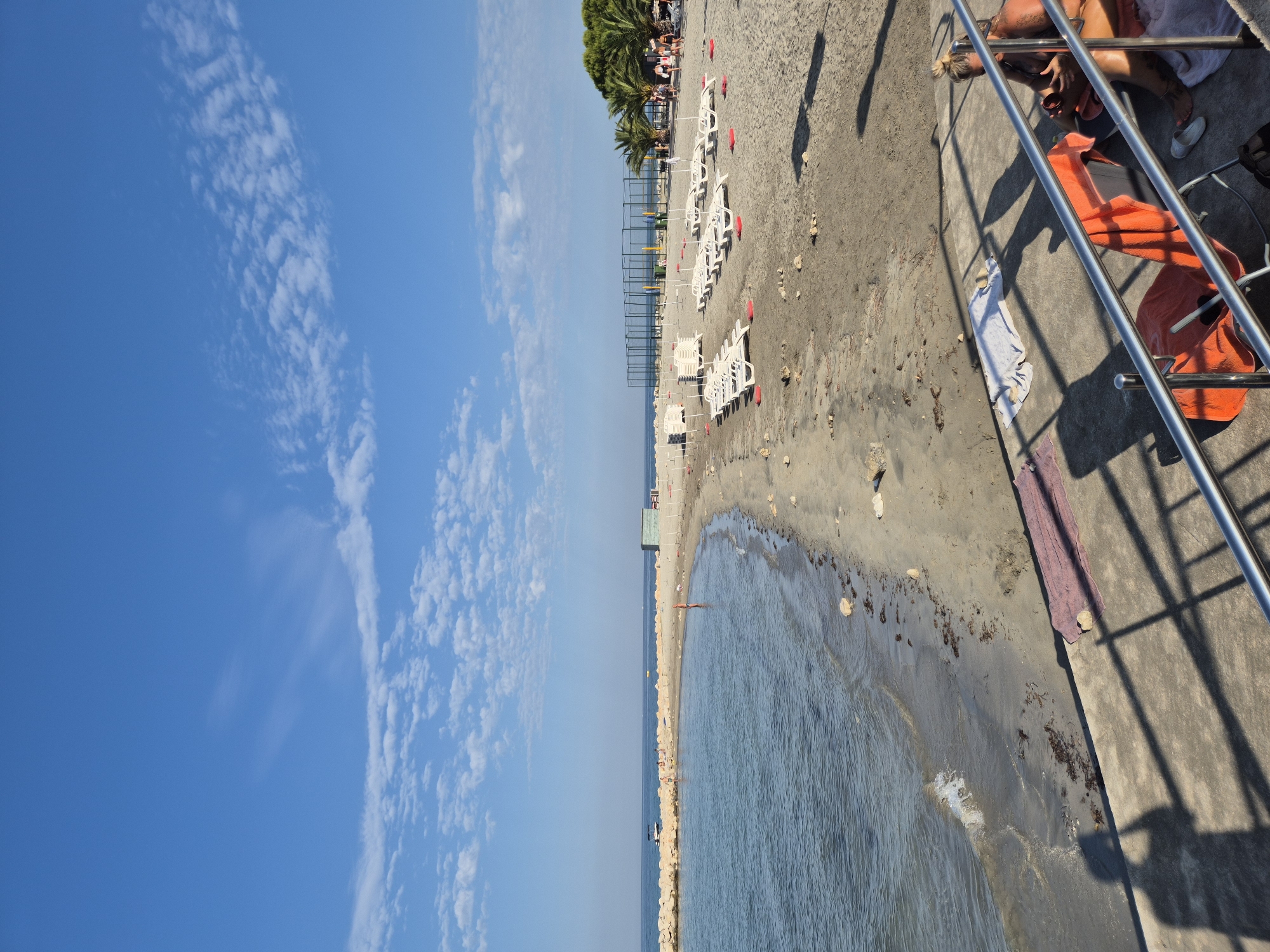 The image shows a beach scene with the sea on the left and a sandy beach on the right. The water is calm with small waves lapping at the shore. The beach is lined with rows of white lounge chairs, some stacked and some set up. There are palm trees and a fenced sports court in the background. Several people are visible on the beach, including some lying on lounge chairs and others walking. In the foreground, there is a concrete walkway with a metal railing, and shadows are cast across it. A person is sitting on the walkway with towels and a chair nearby. The sky is blue with scattered white clouds. The scene appears to be a public beach, possibly a resort, in Monterol, Croatia.
