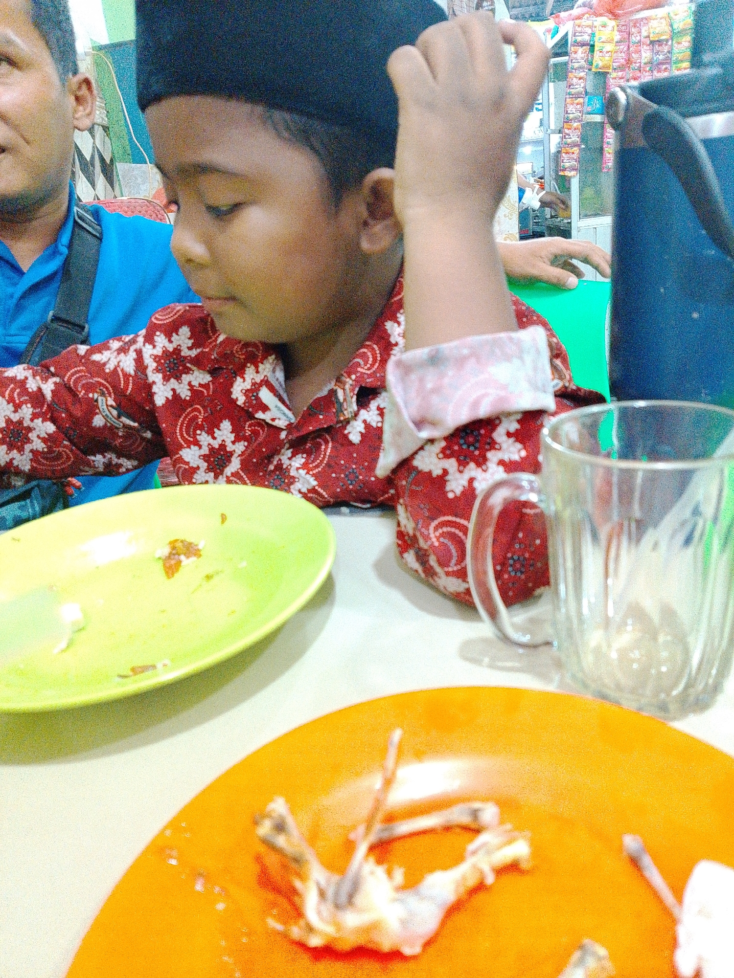 The image depicts a close-up, slightly angled view of two people sitting at a table, likely in a casual eatery in Bandar Setia, Indonesia, as suggested by the context. The foreground is dominated by two plates. The plate on the lower right is bright orange and contains the remnants of food, appearing to be the bones and meat of a small animal, possibly chicken. There are also small white pieces, possibly rice or bread. The plate on the upper left is lime green and has some scattered food particles and a faint smear of sauce. To the right of the green plate is a clear glass mug with a handle, appearing to be empty or containing a small amount of liquid.
The focus of the image shifts to the two individuals. On the left, the shoulder and head of an adult male are visible, wearing a blue shirt and a black strap across his chest, suggesting a bag. He is looking downwards and to the left, his face partially obscured. In the center and to the right, a young boy is the primary subject. He is wearing a red shirt with a white floral pattern and a black peci (a traditional cap). His left arm is raised and bent, with his hand resting on his forehead, giving him a contemplative or possibly tired expression. He is looking down and to the right. His right hand is visible, positioned near his chest.
The background is slightly out of focus but reveals elements of the establishment. Behind the people, shelves stocked with various packaged goods, likely snacks or instant noodles, are visible, suggesting a small shop or warung. A large blue object, possibly a water dispenser or a large container, is situated to the right of the boy. The overall environment appears to be indoors, with artificial lighting contributing to the image's slightly grainy and bright quality. The scene conveys a sense of a meal being concluded or paused.