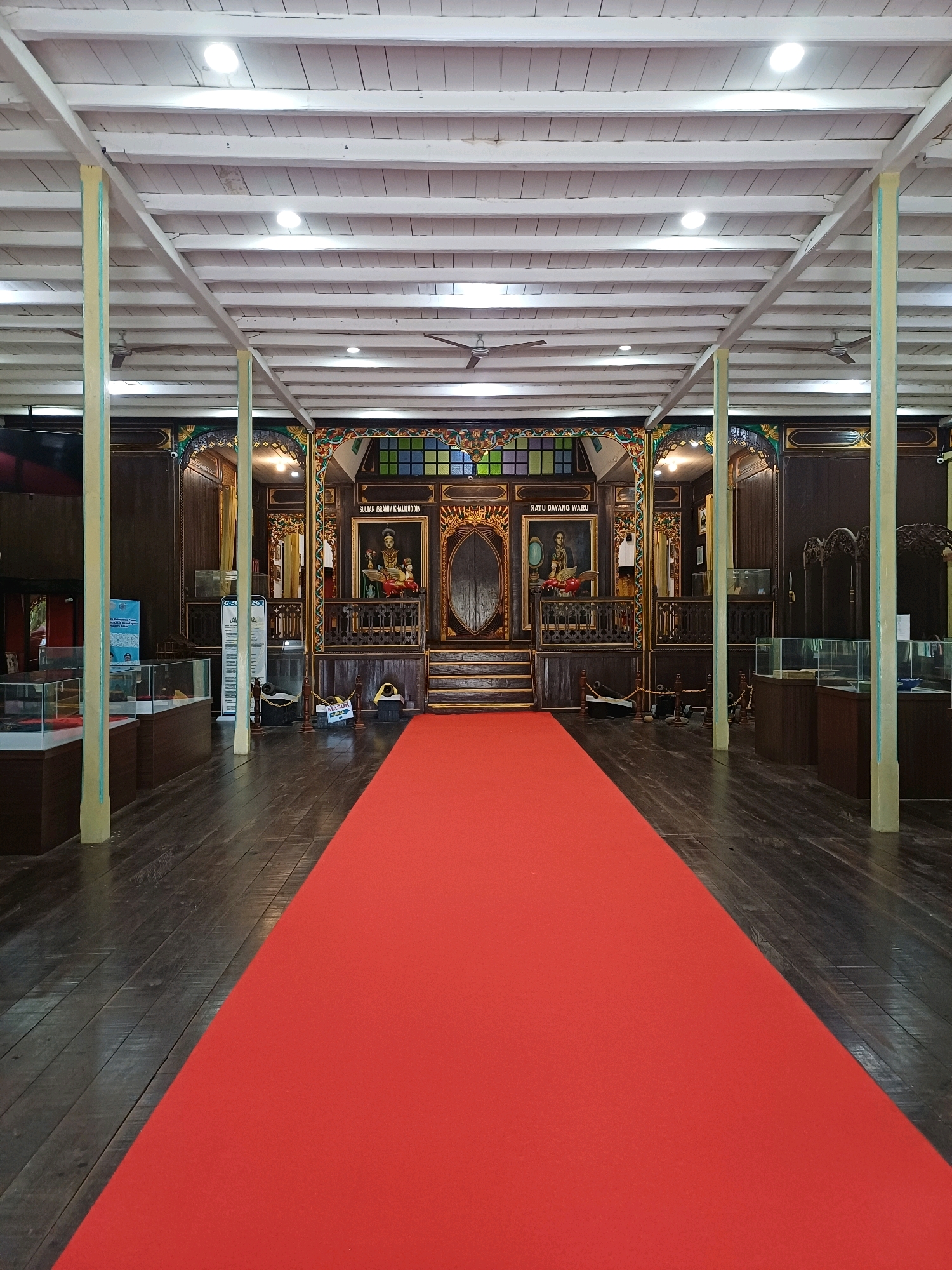 The image shows the interior of a historical building, possibly a museum or a traditional house, located in the city of Pasir Belengkong, Indonesia. The scene is dominated by a bright red carpet laid out on a polished dark wooden floor, leading towards a raised platform at the far end of the room. The platform features ornate wooden carvings and framing, with two large portraits displayed above. The portraits appear to depict figures in traditional attire. The ceiling is high, supported by white-painted beams and industrial-style metal girders, with circular lights and ceiling fans visible.
Along the sides of the room, there are glass display cases containing what seem to be artifacts, suggesting a museum setting. The walls are panelled with dark wood, decorated with intricate carvings and colorful stained glass windows above the portraits. Two large, slender pillars with light yellow and light blue accents are positioned in the foreground, partially obscuring the view of the display cases. The overall ambiance suggests a place of cultural or historical significance, prepared for visitors or an event. There are no people visible in the image.