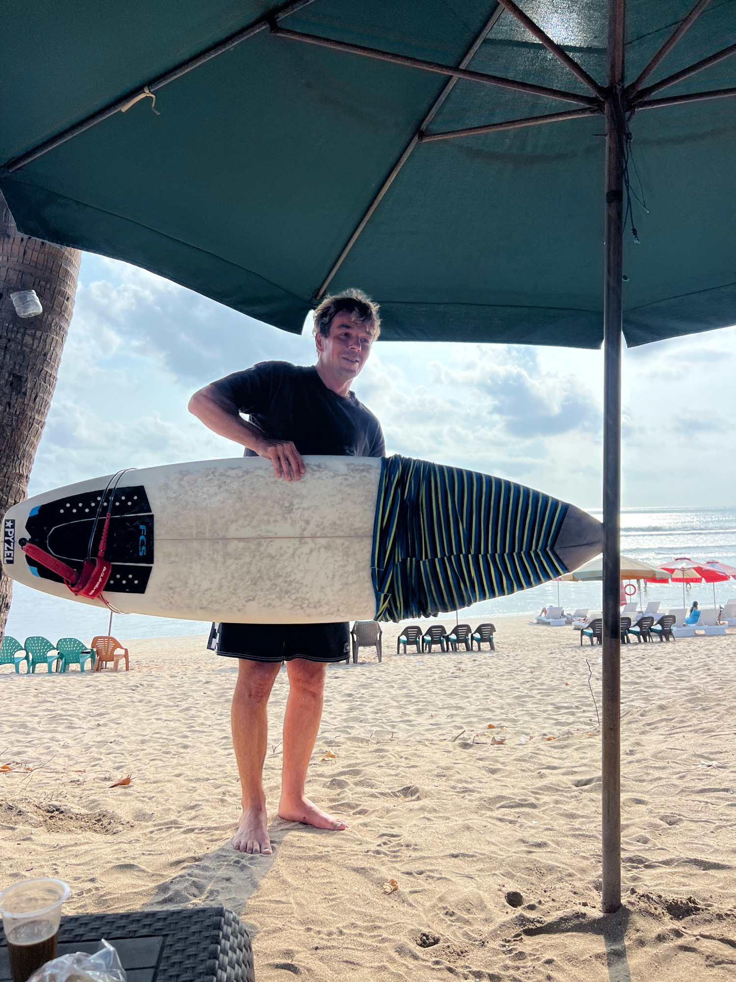 A man stands on a sandy beach in Kuta, Indonesia, holding a surfboard. He is wearing a black t-shirt and black shorts, with his feet bare. The surfboard is white with a black traction pad and a red leash. The surfboard is partially covered by a blue and yellow striped fabric. Behind him, the ocean stretches to the horizon, with a clear sky and scattered clouds. Beach chairs and umbrellas are visible in the background, along with a palm tree to the left of the frame. The scene is illuminated by natural sunlight.