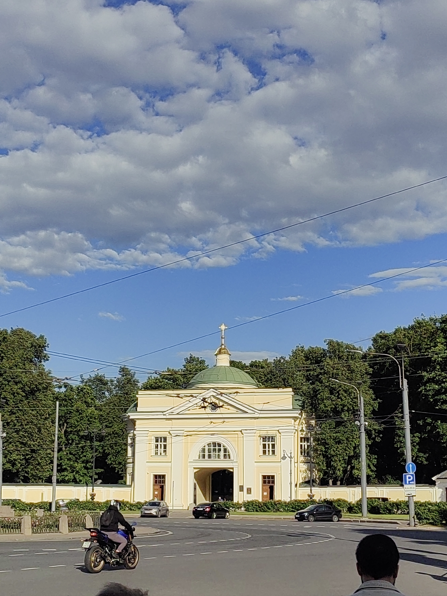 A sunny day in Saint Petersburg, Russia, features a yellow building with a green dome and a cross on top, likely a church or religious institution. The building is framed by lush green trees on both sides. In the foreground, a person rides a motorcycle on a street with white road markings. Several cars are visible on the road, and a blue and white parking sign is posted on the right side of the image. The sky is a vibrant blue with scattered white clouds. The scene suggests a typical urban environment in Saint Petersburg during the daytime.