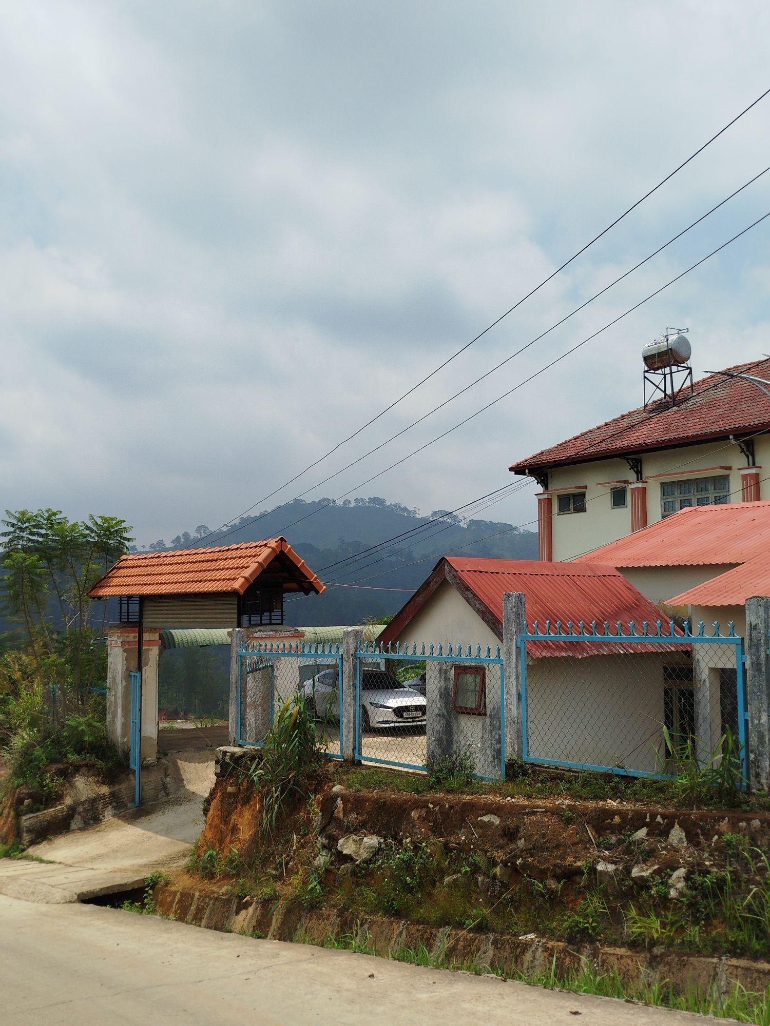 The image presents a scene in Đa Nghịt 1, Vietnam, featuring a building complex with a mix of architectural styles. The building on the right has a multi-tiered red tile roof and cream-colored walls, with a water tank visible on the roof. Adjacent is a smaller, single-story structure, also with a red tile roof. In front, a faded blue metal fence runs along the perimeter, slightly overgrown with vegetation.
To the left stands a distinctive, elevated red-roofed structure, possibly a guard house or viewpoint, accessed by a concrete path leading up from the foreground. The landscape in the background includes rolling hills covered in trees, partially obscured by an overcast sky. Overhead, power lines stretch across the frame. A car is partially visible through the fencing. The foreground shows a concrete path and a dirt embankment with scattered vegetation.